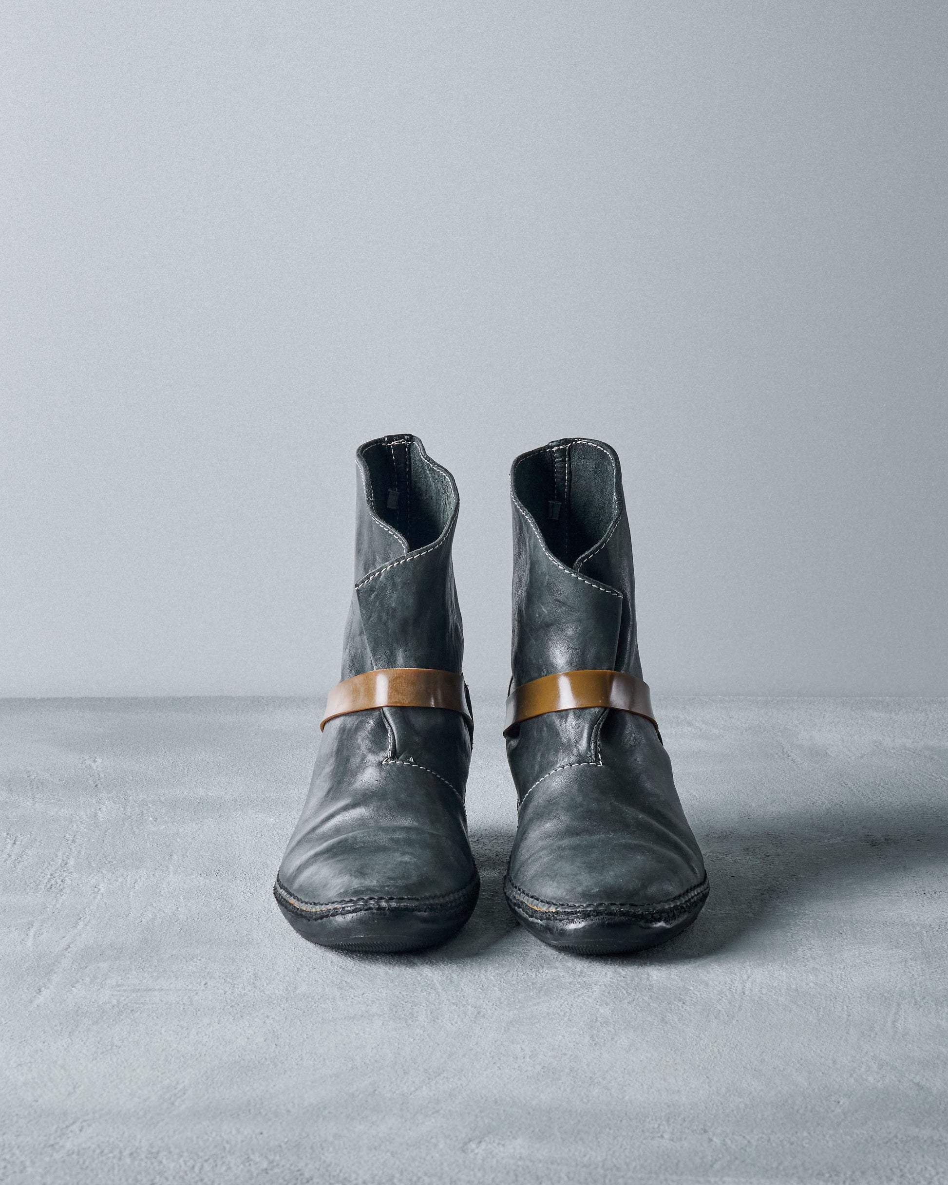 Black leather Carol Christian Poell lace-up boots with a thick rubber band wrapping around the ankle, positioned against a grey background.