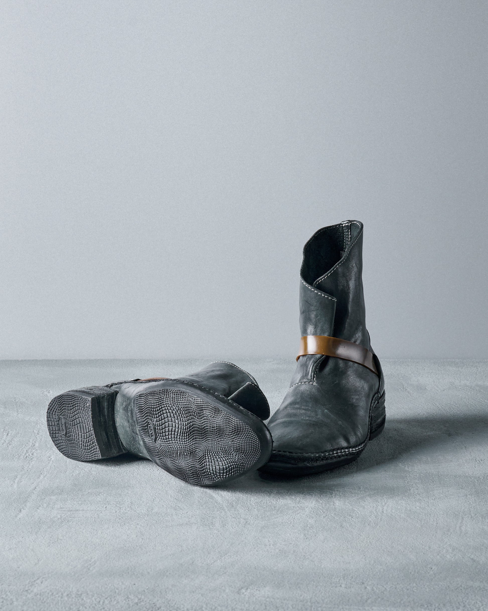 A pair of worn black leather Carol Christian Poell cowboy boots, one standing upright and the other lying on its side, showcasing a textured sole. The setting is a plain gray surface, conveying a rustic and rugged tone.