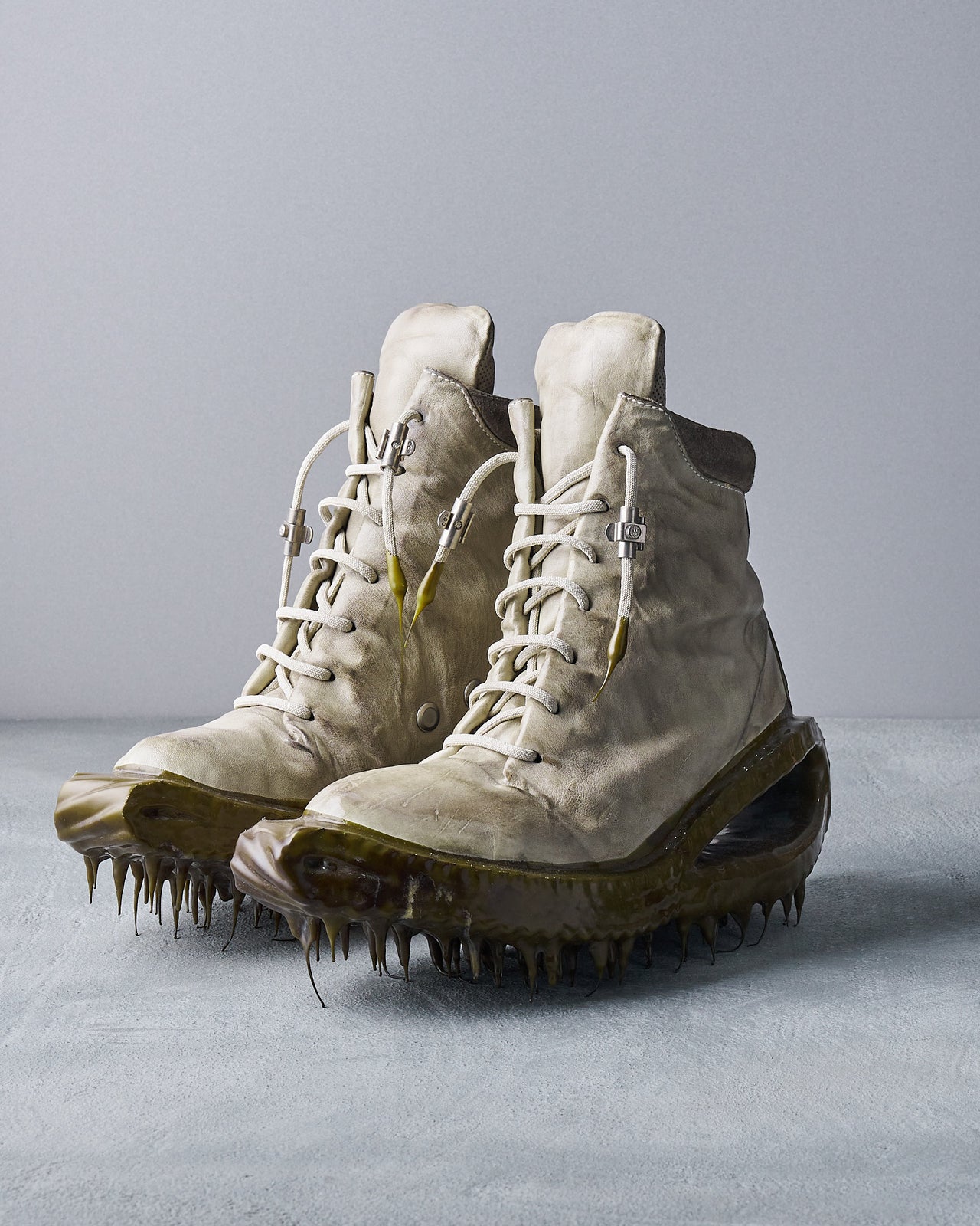 Worn, white high-top Carol Christian Poell sneakers with thick, translucent, spike-covered soles on a plain grey surface. The tone is mysterious and surreal.