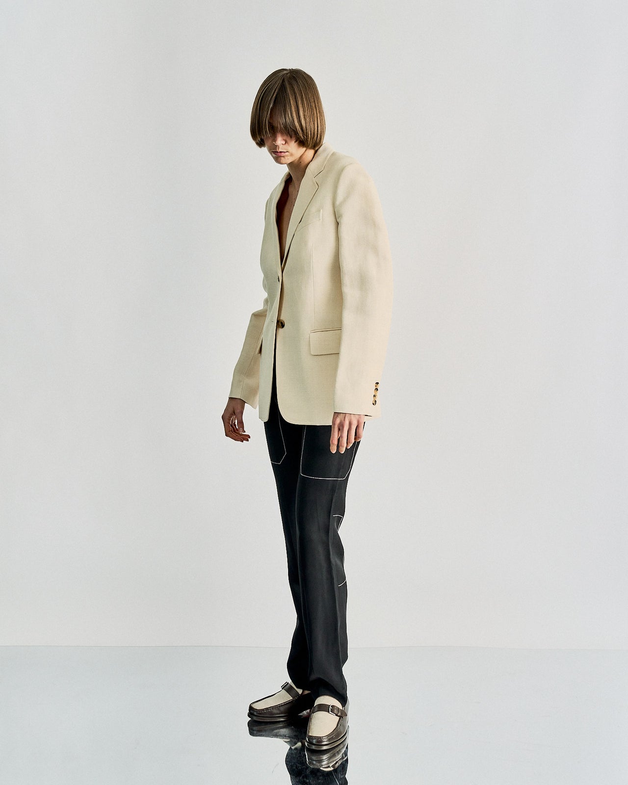 A person with a short bob hairstyle wears a beige Celine blazer, black pants with white stitching, and two-tone loafers. Their head is slightly bowed, creating a contemplative tone.