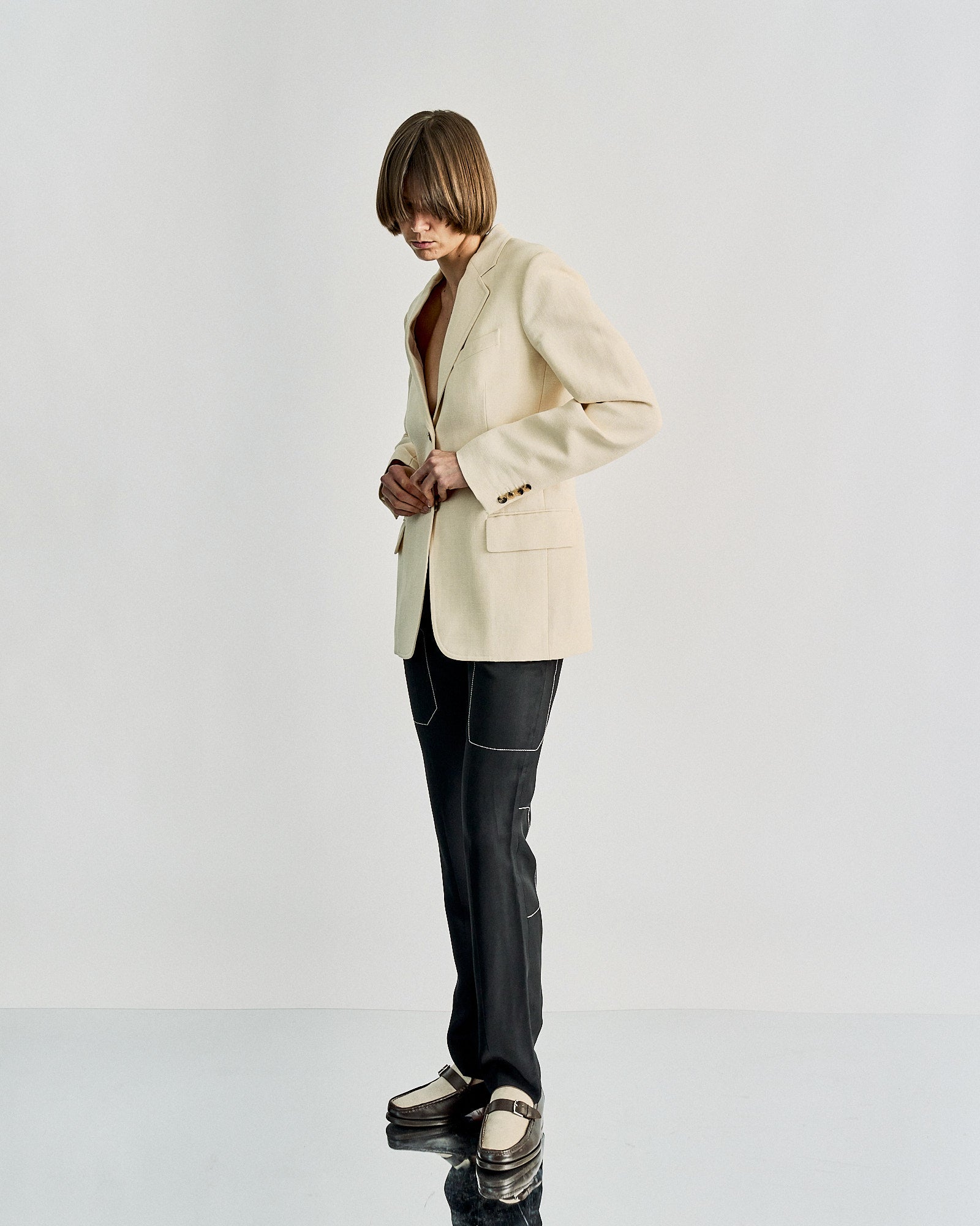 A person stands against a white background wearing a cream Celine blazer, dark pants with white stitching, and loafers. The tone is modern and stylish.
