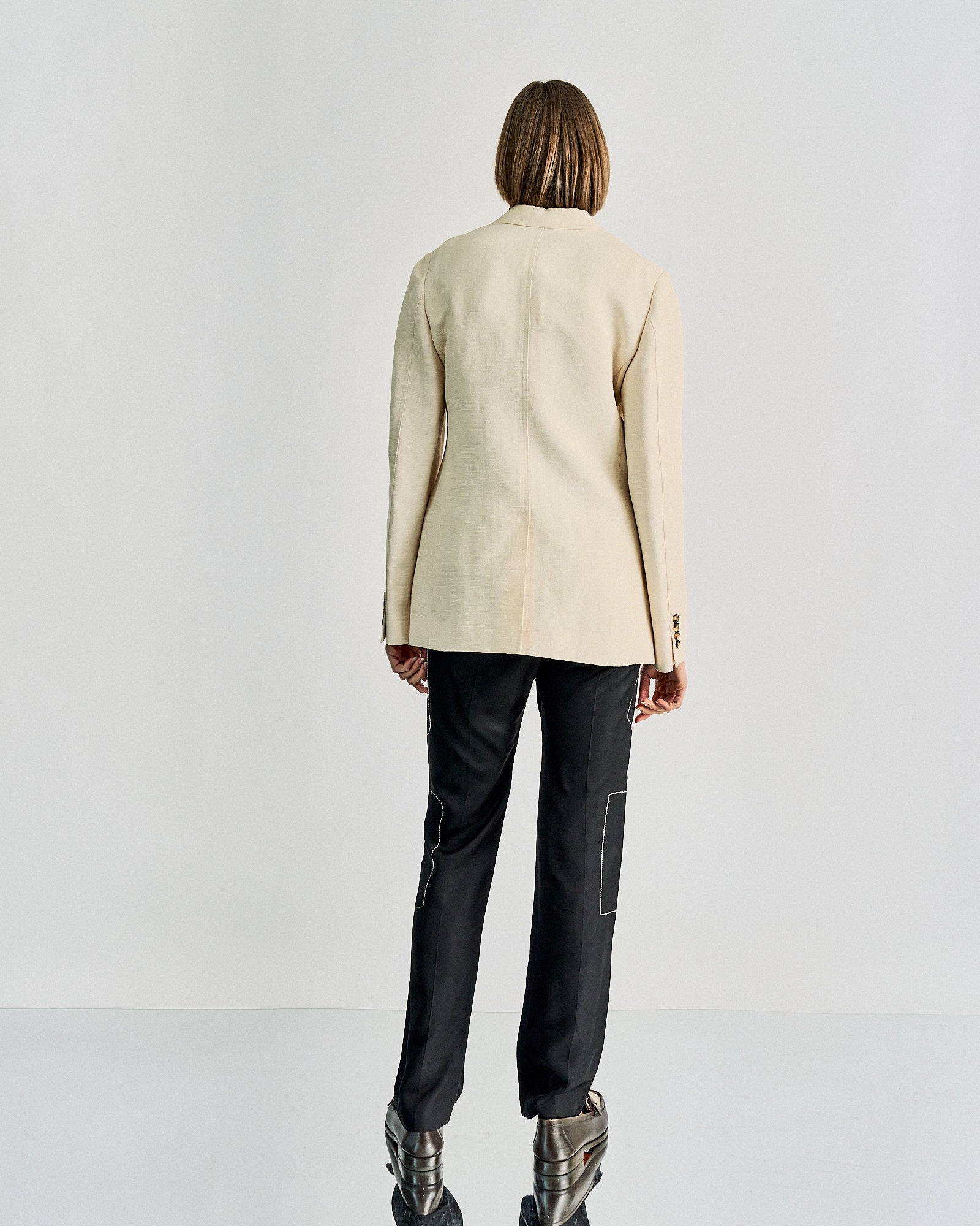 Person with brown bob haircut wearing a beige Celine blazer, black pants with white stitching, and metallic shoes, standing against a neutral background.