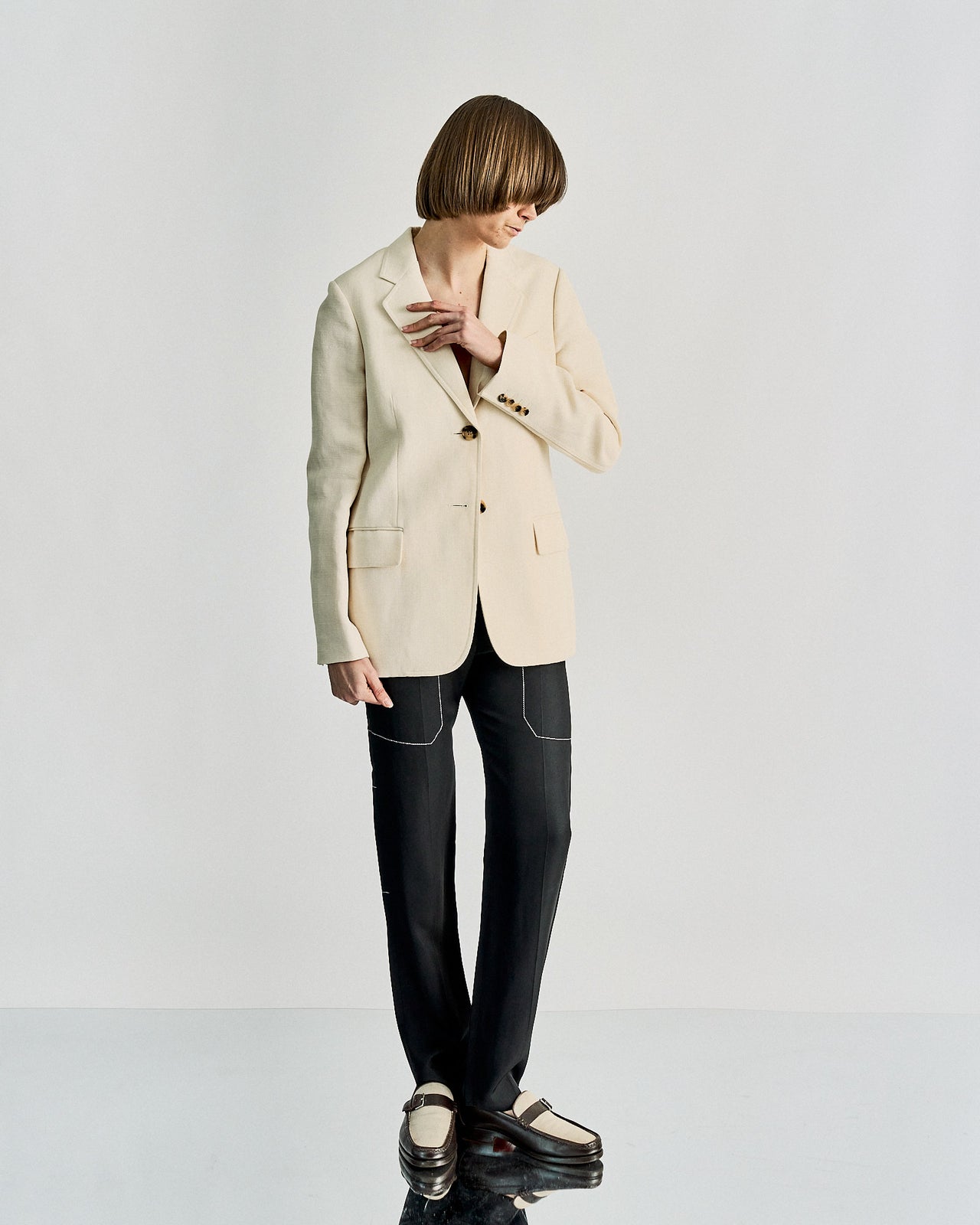 A person with a short bob hairstyle wears a beige Celine blazer, black pants with white stitching, and two-tone loafers. Their head is slightly bowed, creating a contemplative tone.