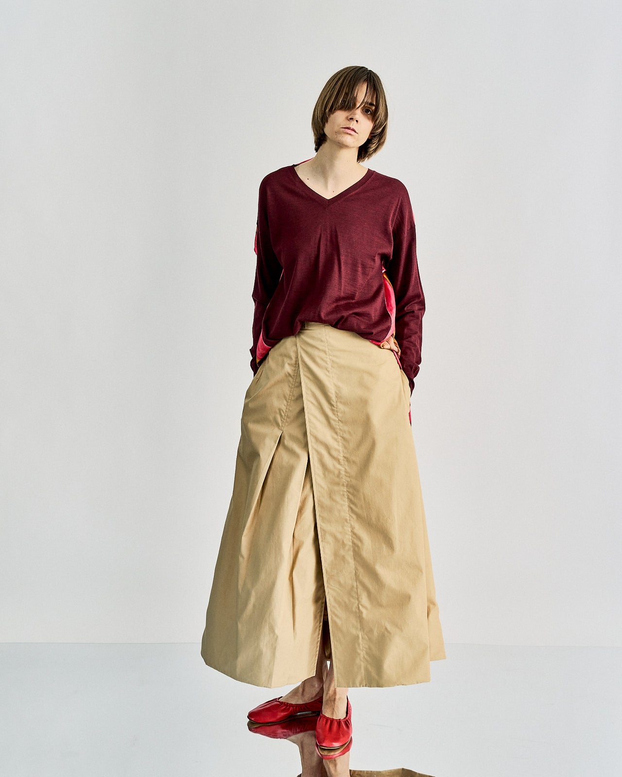 A person stands facing away, showcasing a vibrant, abstract-patterned Celine scarf top in pink and brown over a burgundy shirt. They wear a beige skirt and pink shoes.