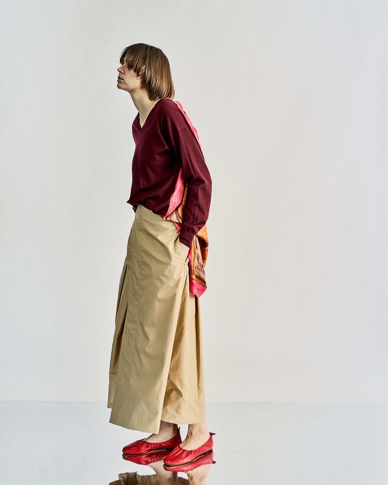 Person in profile wearing a burgundy Celine top and beige wide-leg pants, with a colourful patterned scarf and red shoes. The pose is thoughtful and casual.