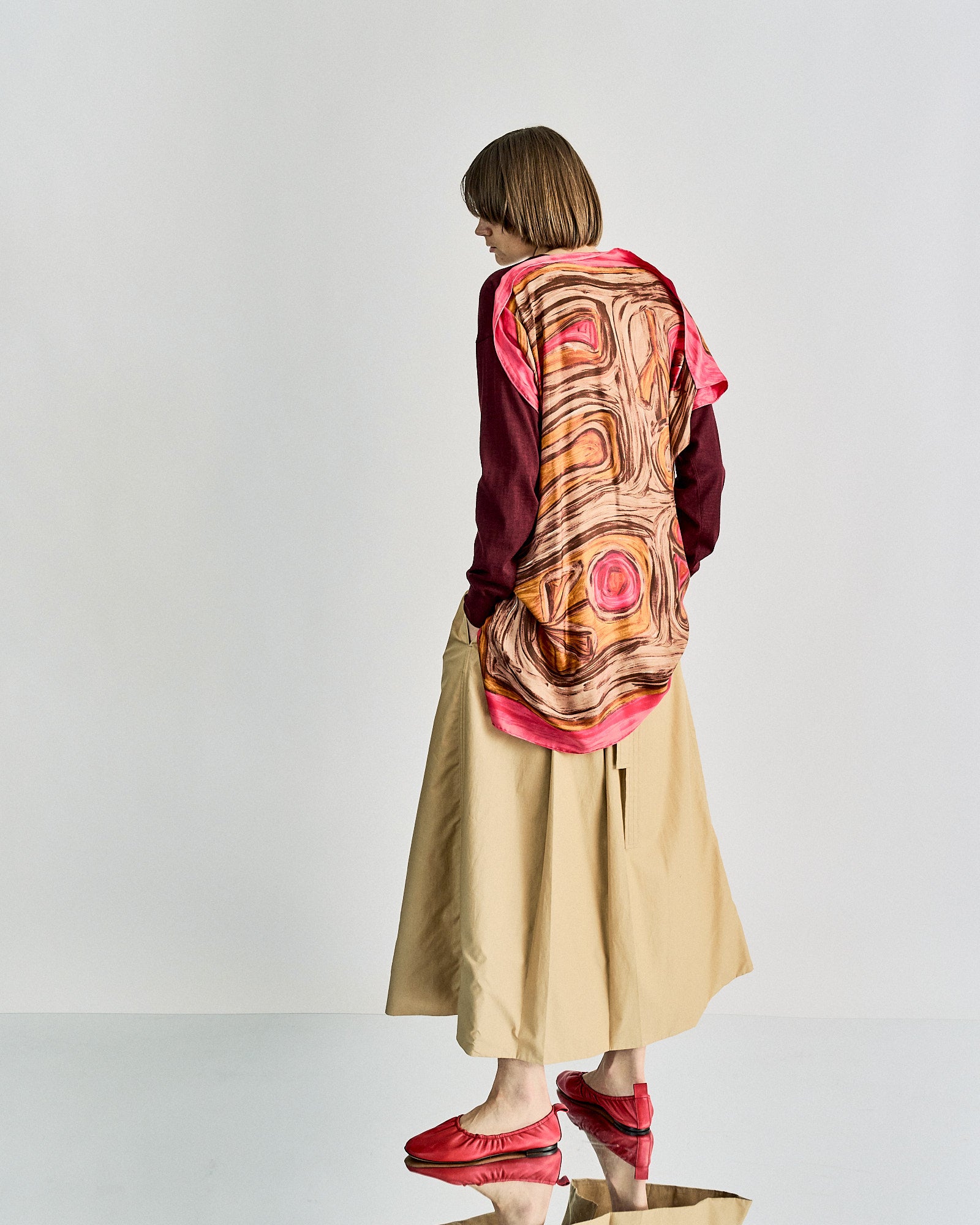 Person in profile wearing a burgundy Celine top and beige wide-leg pants, with a colourful patterned scarf and red shoes.