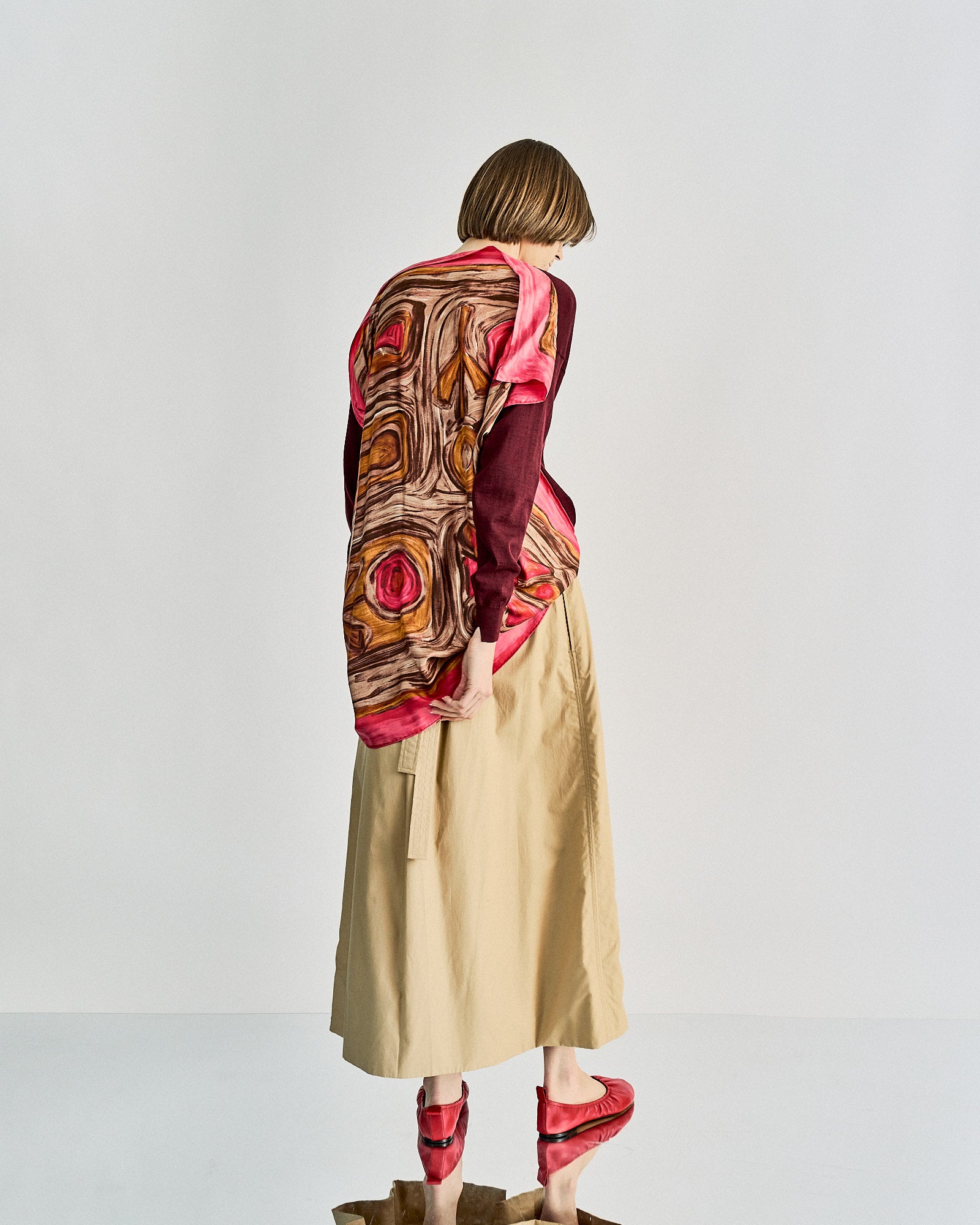 A model in a beige skirt and burgundy Celine top is draped with a colourful scarf featuring pink and brown patterns. Her red shoes match the vibrant scarf.