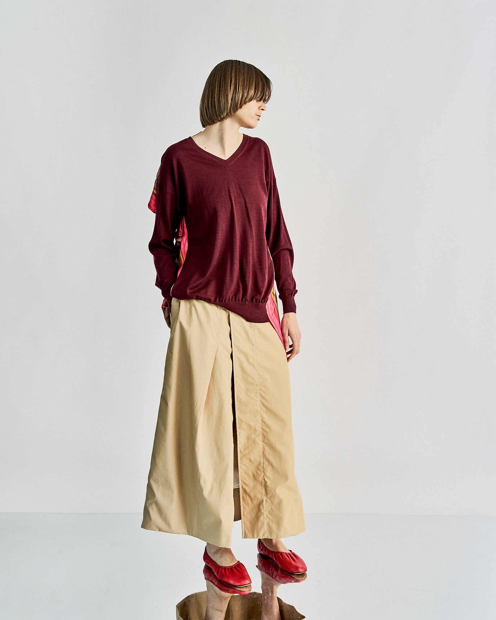 A person wearing a maroon Celine sweater and beige wide-leg pants stands against a plain background. Red shoes add a pop of colour.