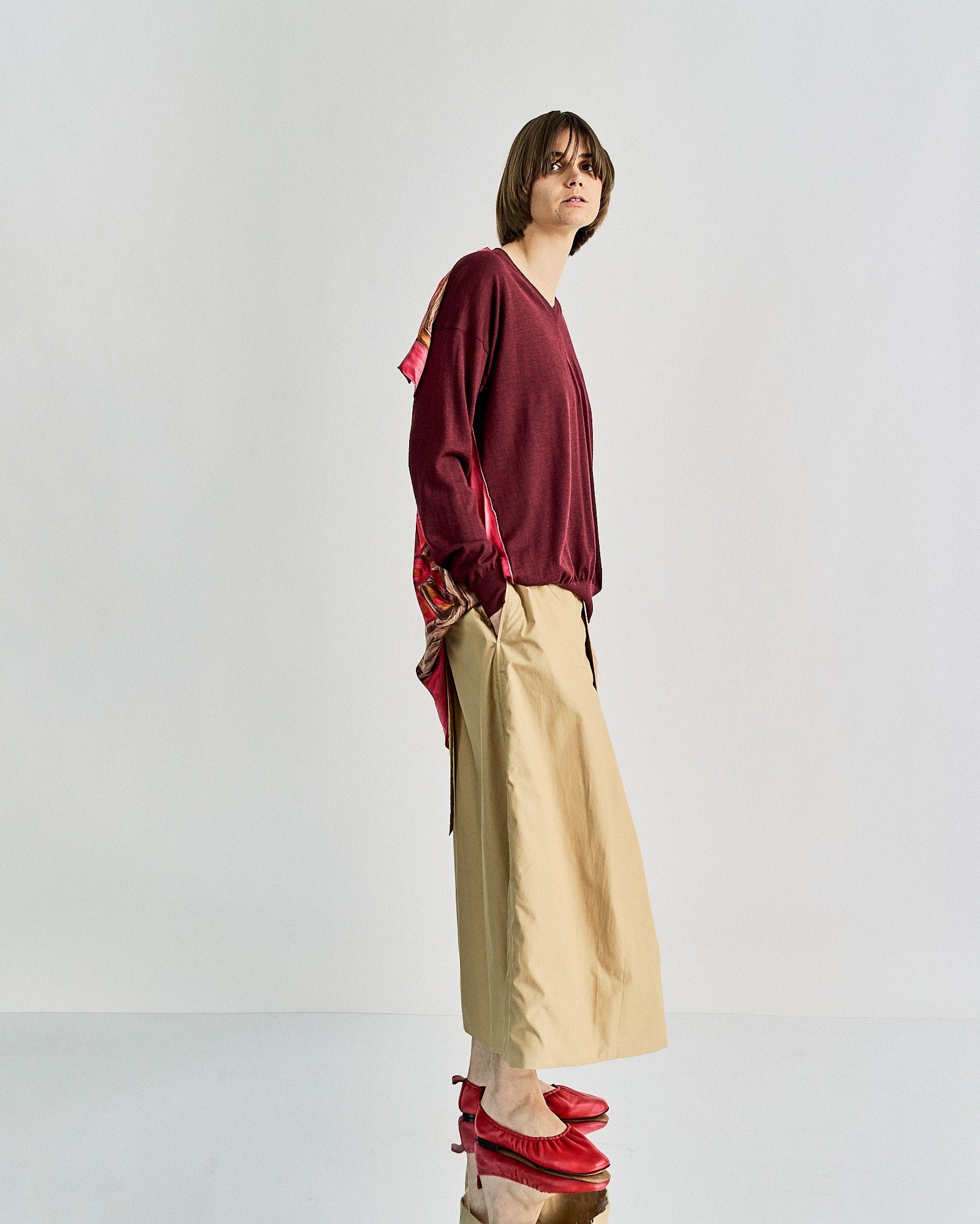 A person stands in a neutral pose wearing a burgundy Celine sweater with patterned accents, beige wide-legged pants, and red shoes. The background is plain white.