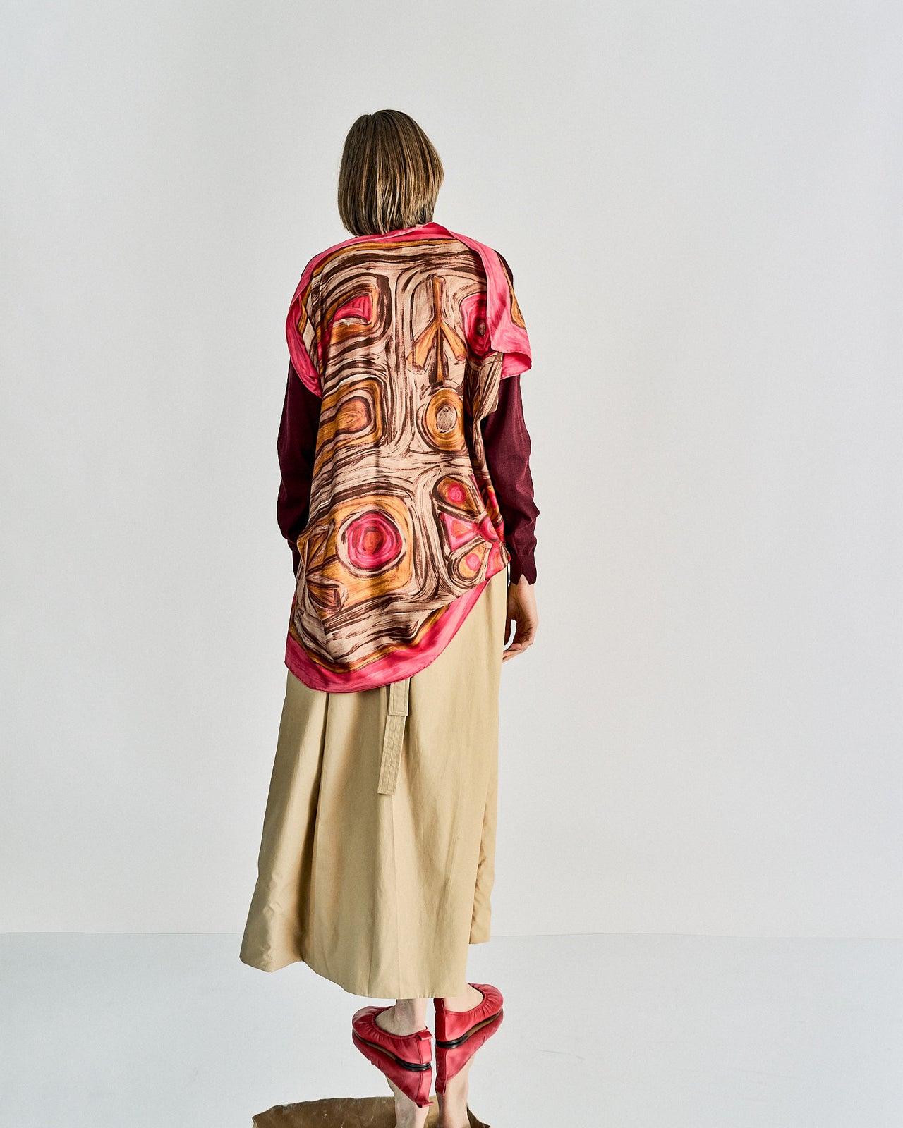 A person stands facing away, showcasing a vibrant, abstract-patterned Celine scarf top in pink and brown over a burgundy shirt. They wear a beige skirt and pink shoes.