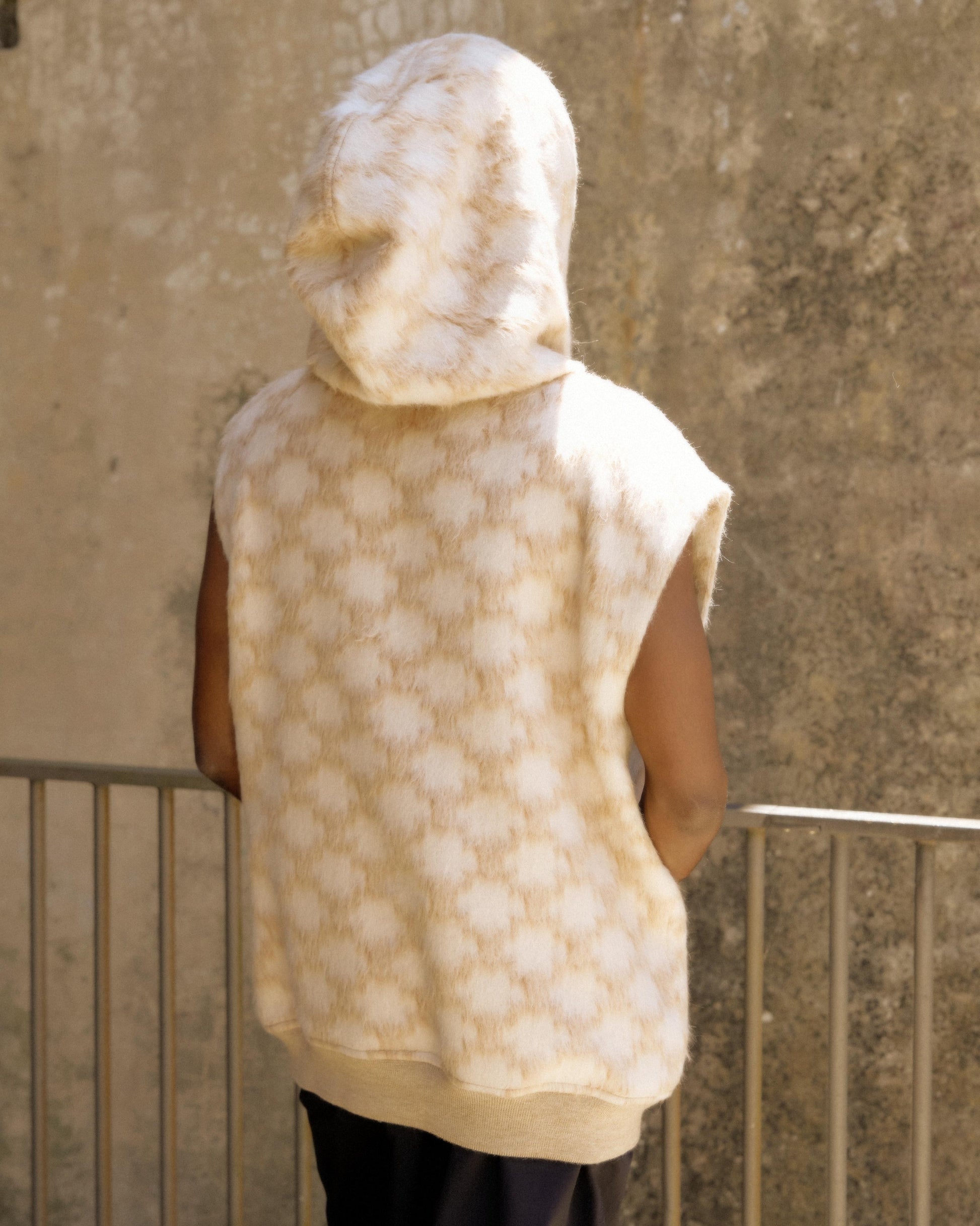A person stands with their back to the camera, wearing a sleeveless, hooded beige Celine top with a soft, textured pattern. The background is a textured wall.