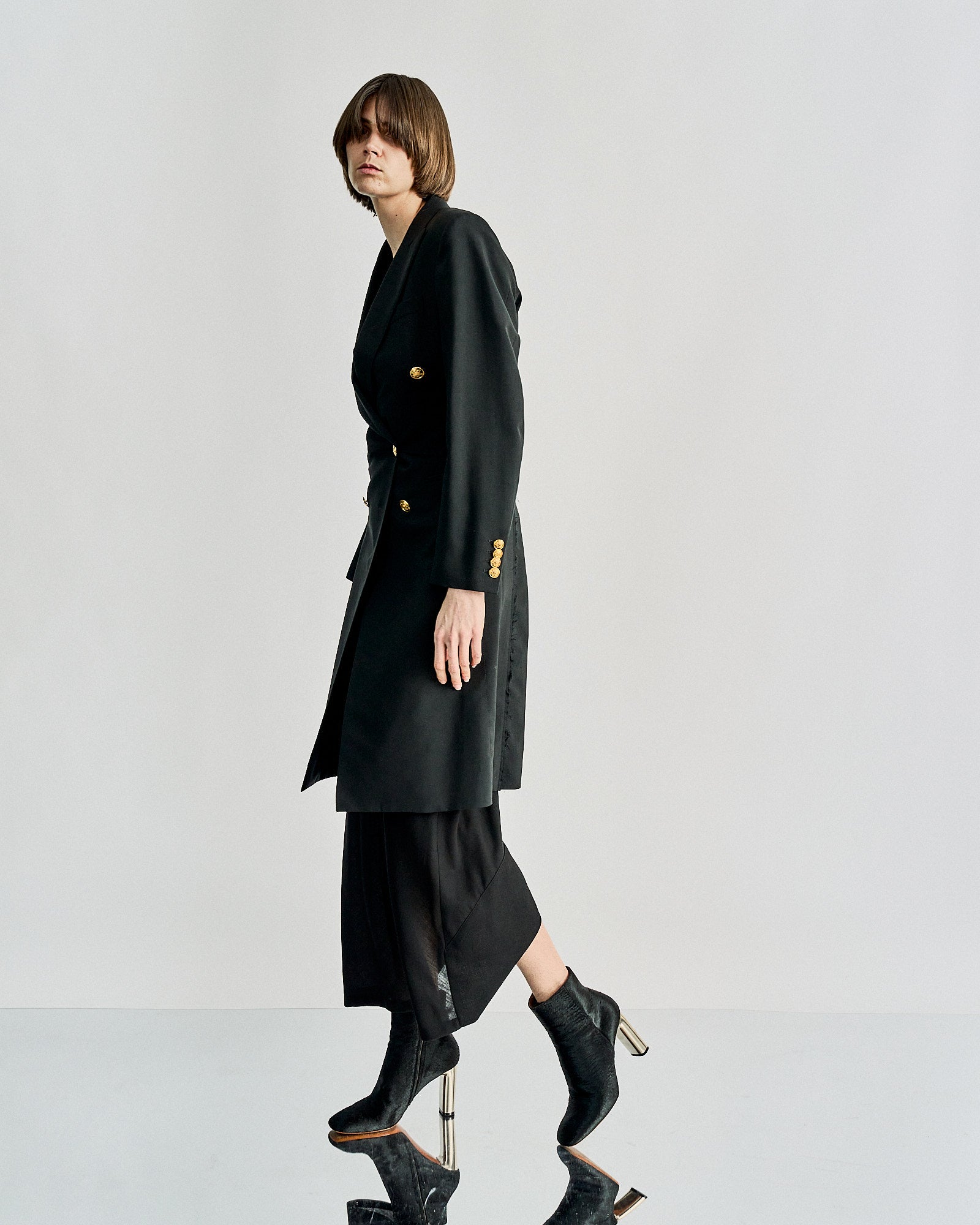 A woman in a sleek black Celine coat with gold buttons and heeled boots walks confidently against a neutral background, exuding elegance and modern fashion.