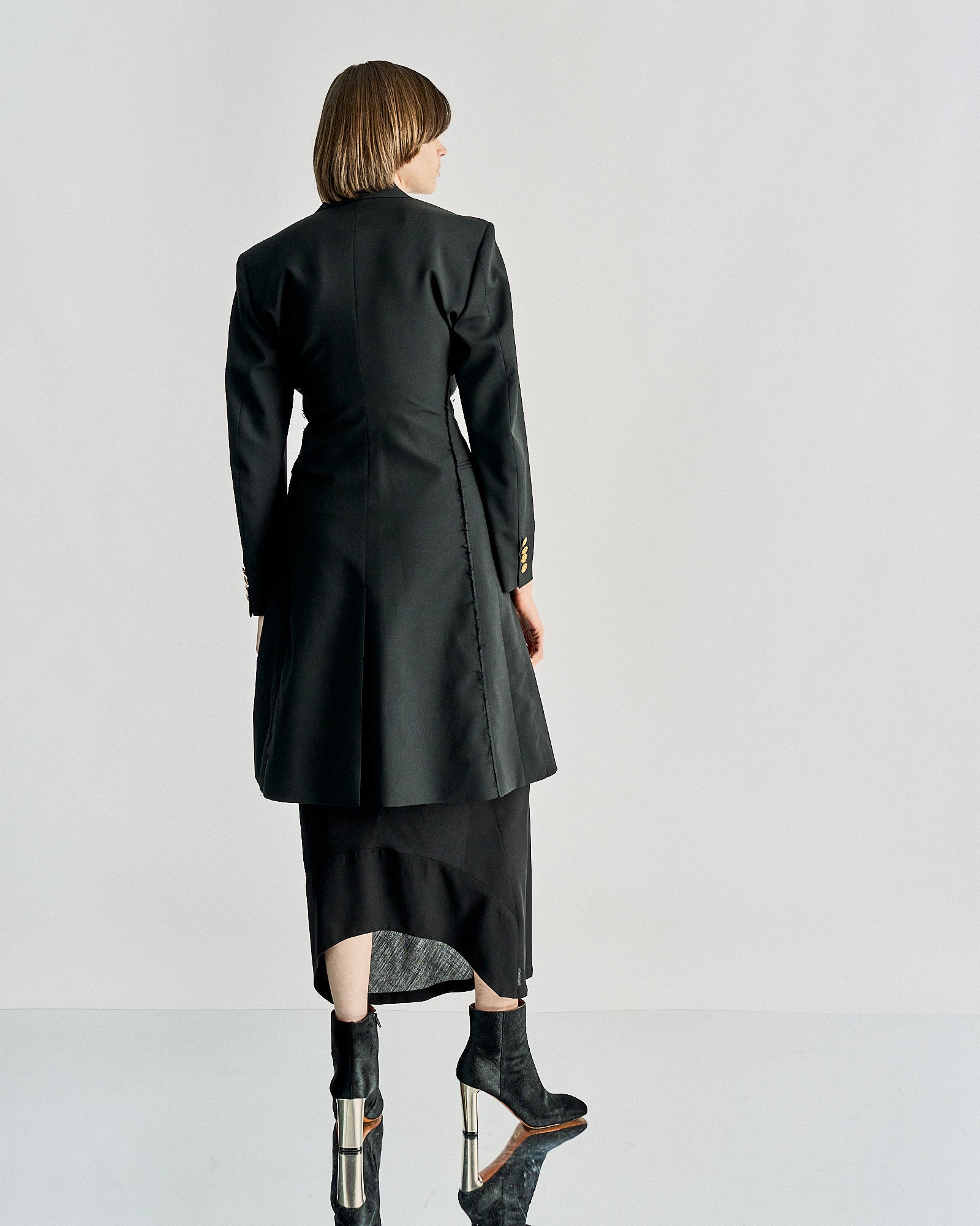A person with short hair faces away, wearing a long black Celine coat over a black skirt and boots. The sleek, modern outfit exudes a confident, stylish tone.