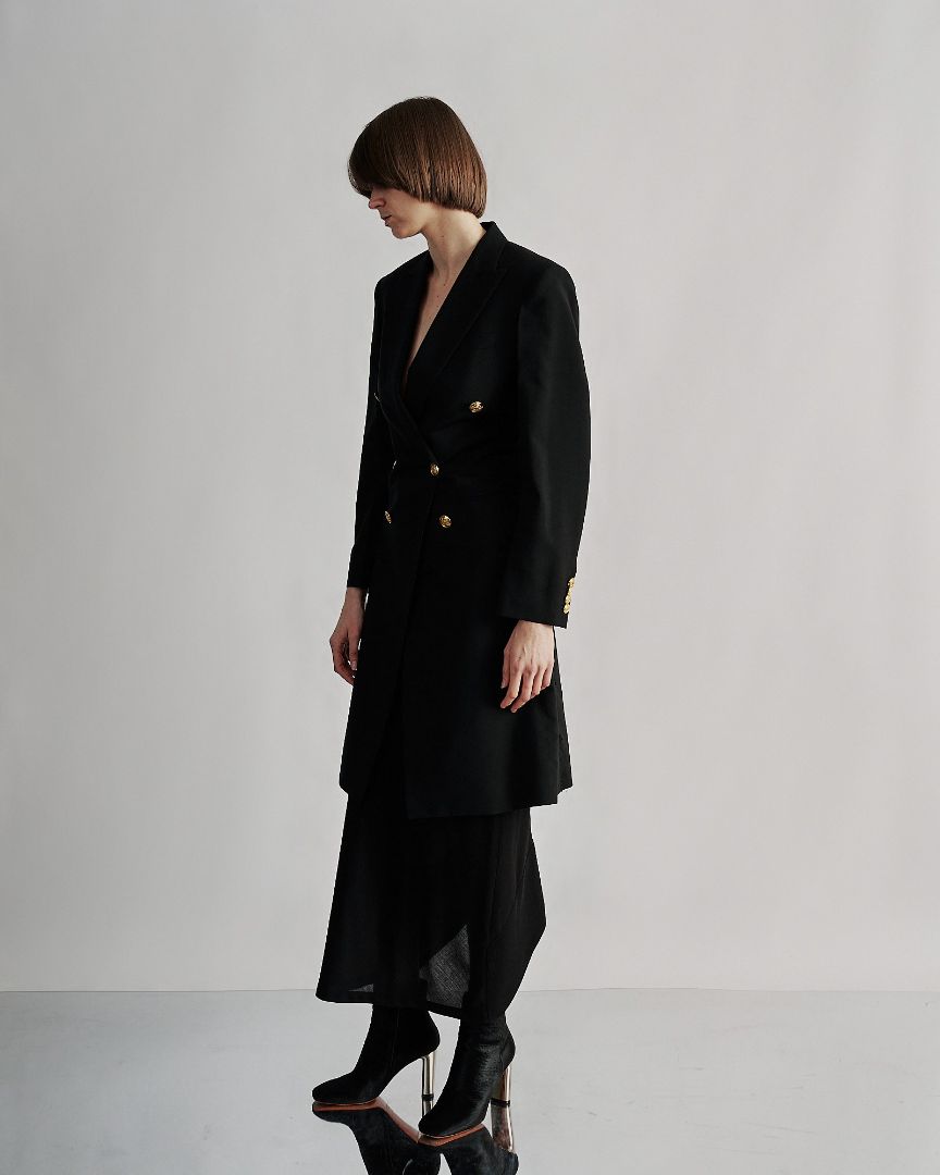 Woman in a black tailored Celine coat with gold buttons, stands in profile against a neutral background. Her short hair complements the modern, elegant look.