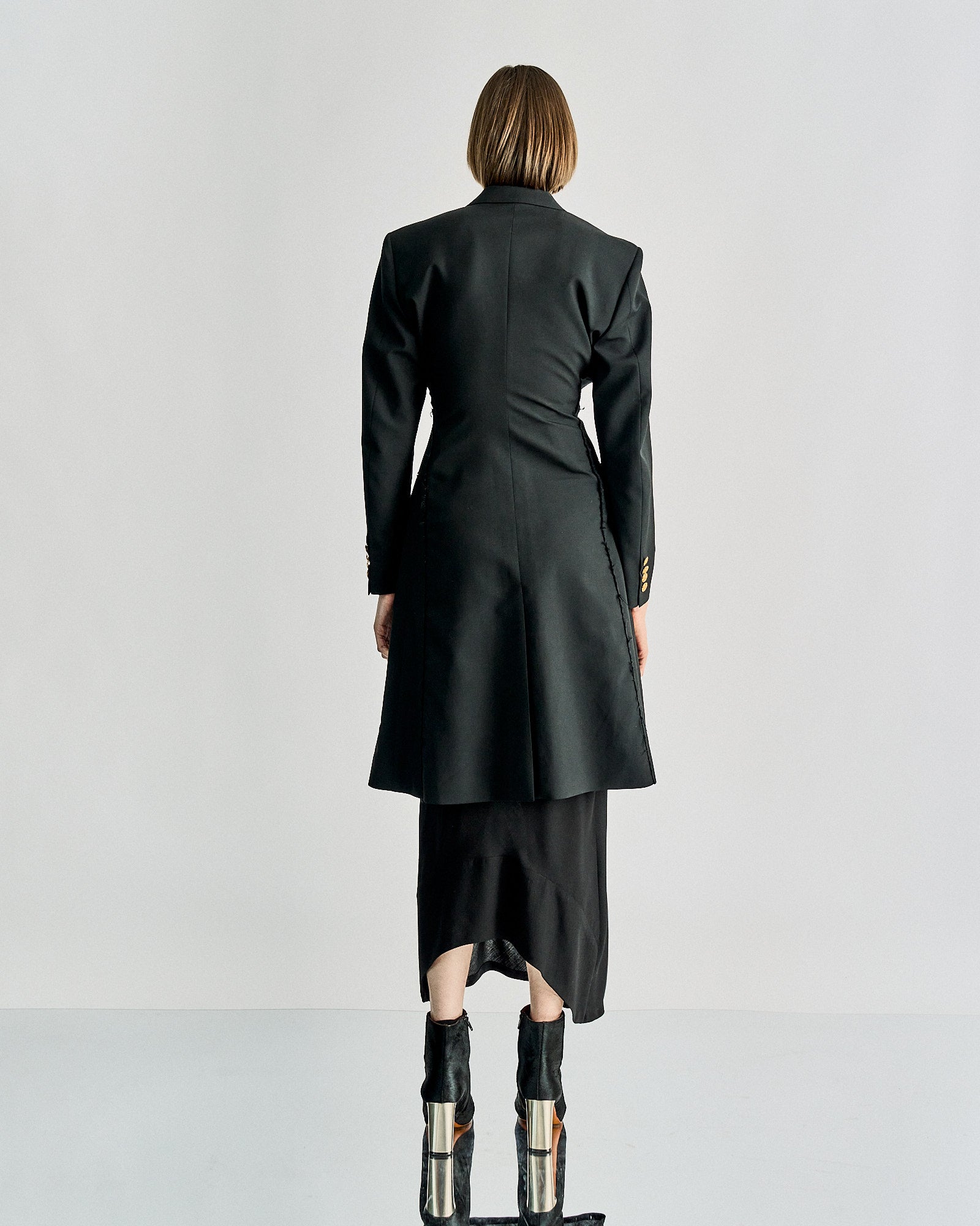 A person stands with their back to the camera, wearing a long black Celine coat over a black dress. They have short brown hair and wear high black boots.
