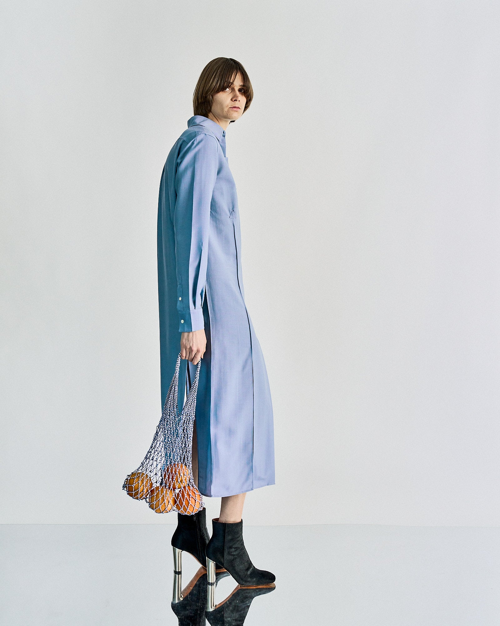A person in a long, light blue Celine dress stands sideways, holding a mesh bag of oranges. They wear black heeled boots, conveying a modern, stylish vibe.