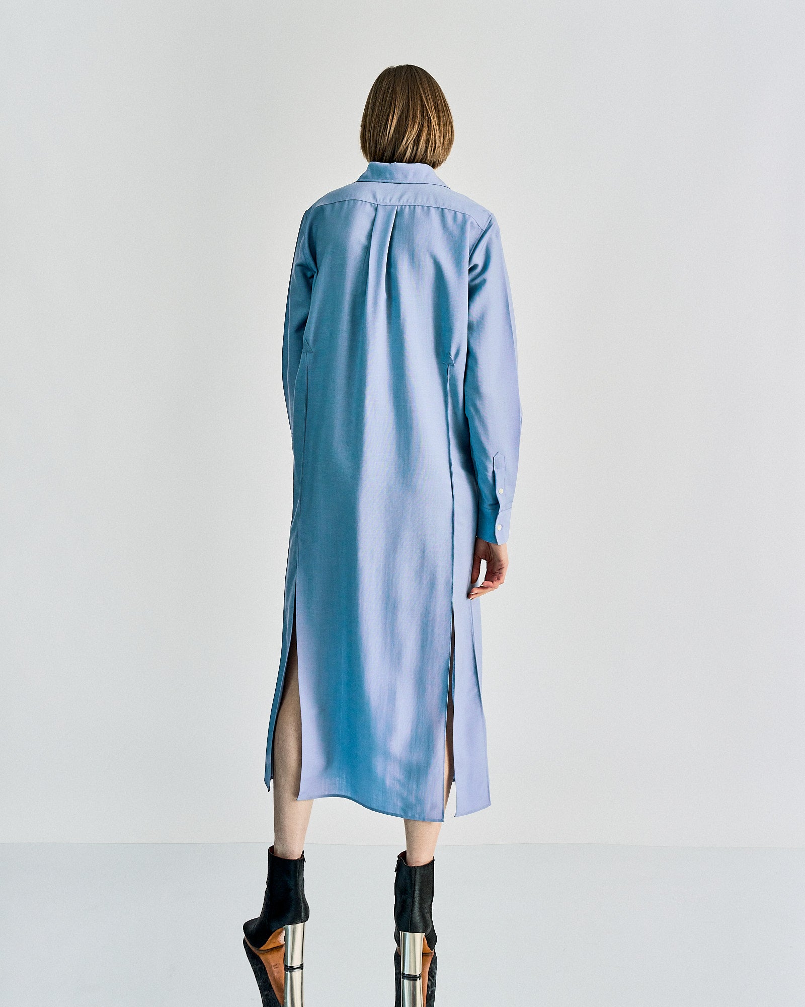 Woman with short hair in a long, light blue Celine shirt dress, standing on a reflective surface. She wears black and brown heeled boots, image feels modern and minimalist.