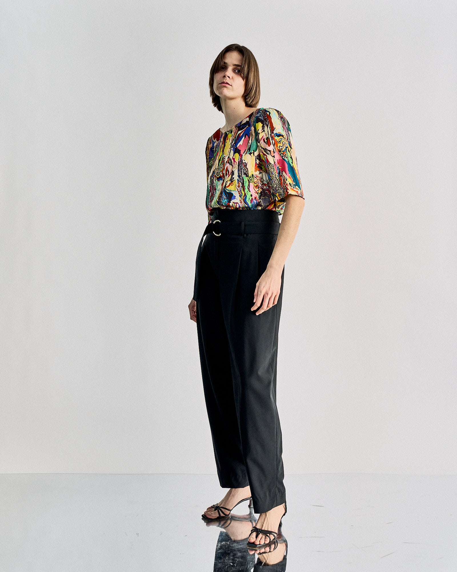 A person in a colourful abstract patterned blouse and high-waisted black Celine pants stands confidently in an empty space, exuding a modern, stylish vibe.
