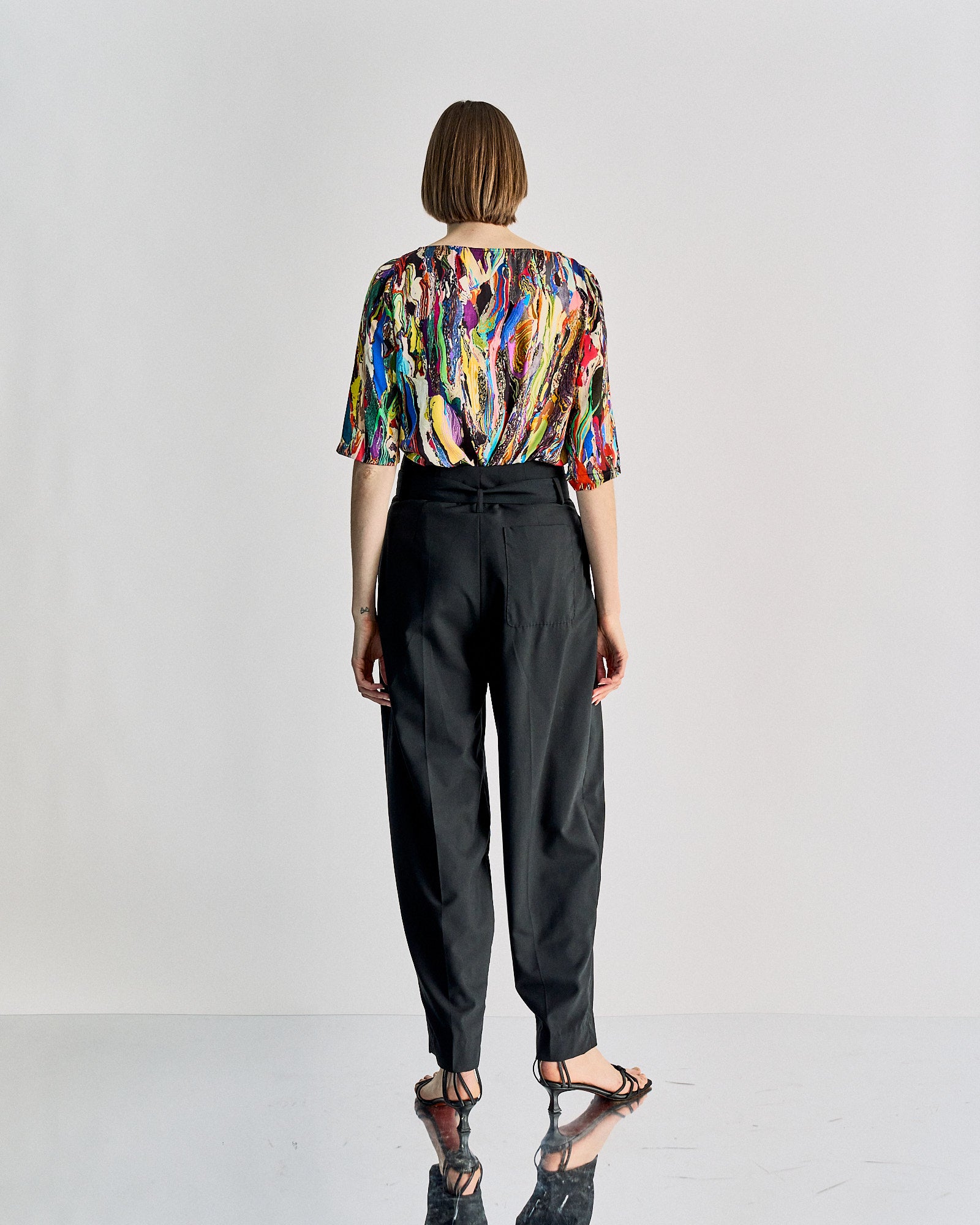 A person stands with their back to the camera, wearing a vibrant, abstract-patterned blouse and black high-waisted Celine trousers, exuding a modern, artistic vibe.