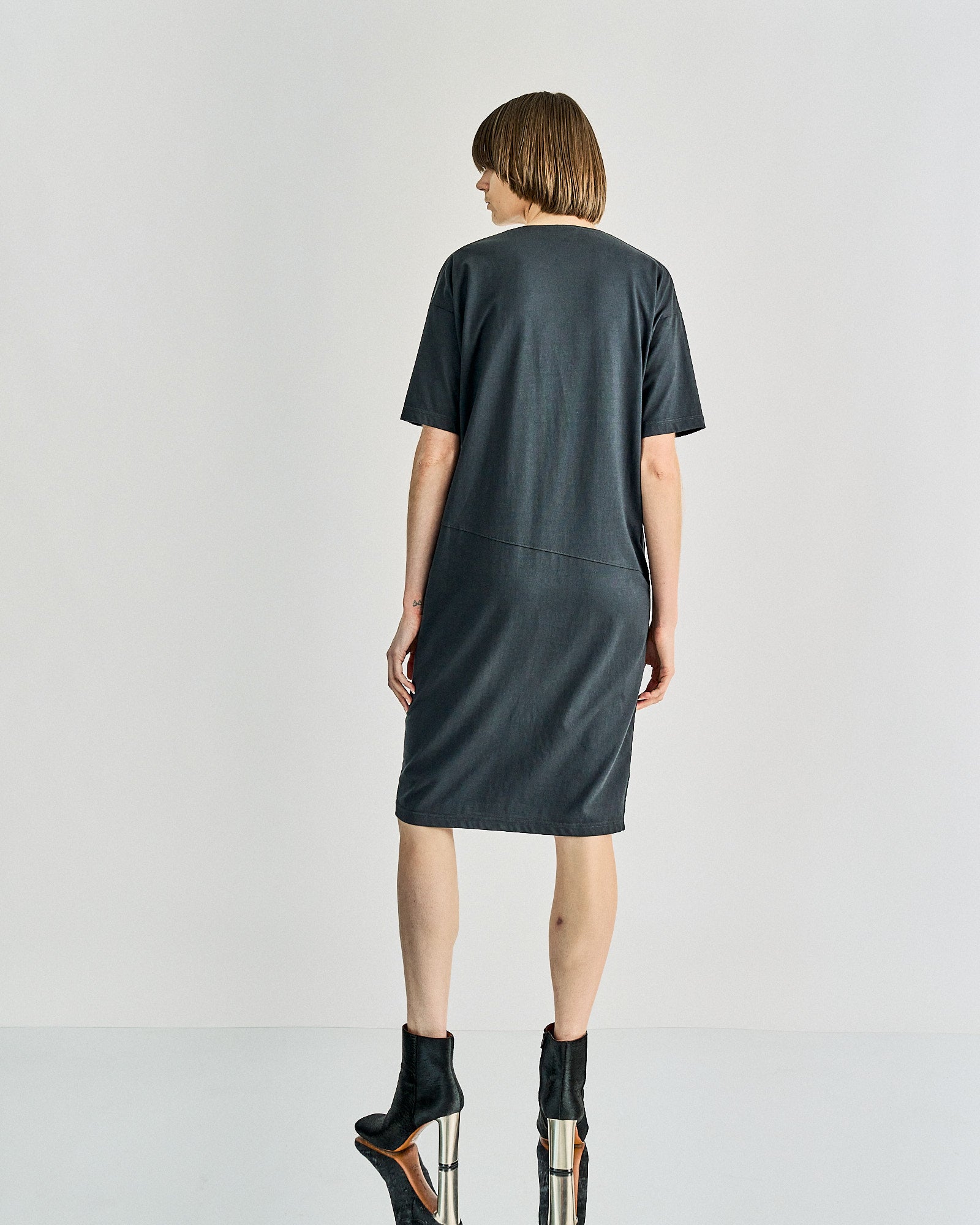A person with short brown hair stands with their back to the camera, wearing a simple, dark gray Celine dress and black ankle boots, creating a minimalistic and modern vibe.
