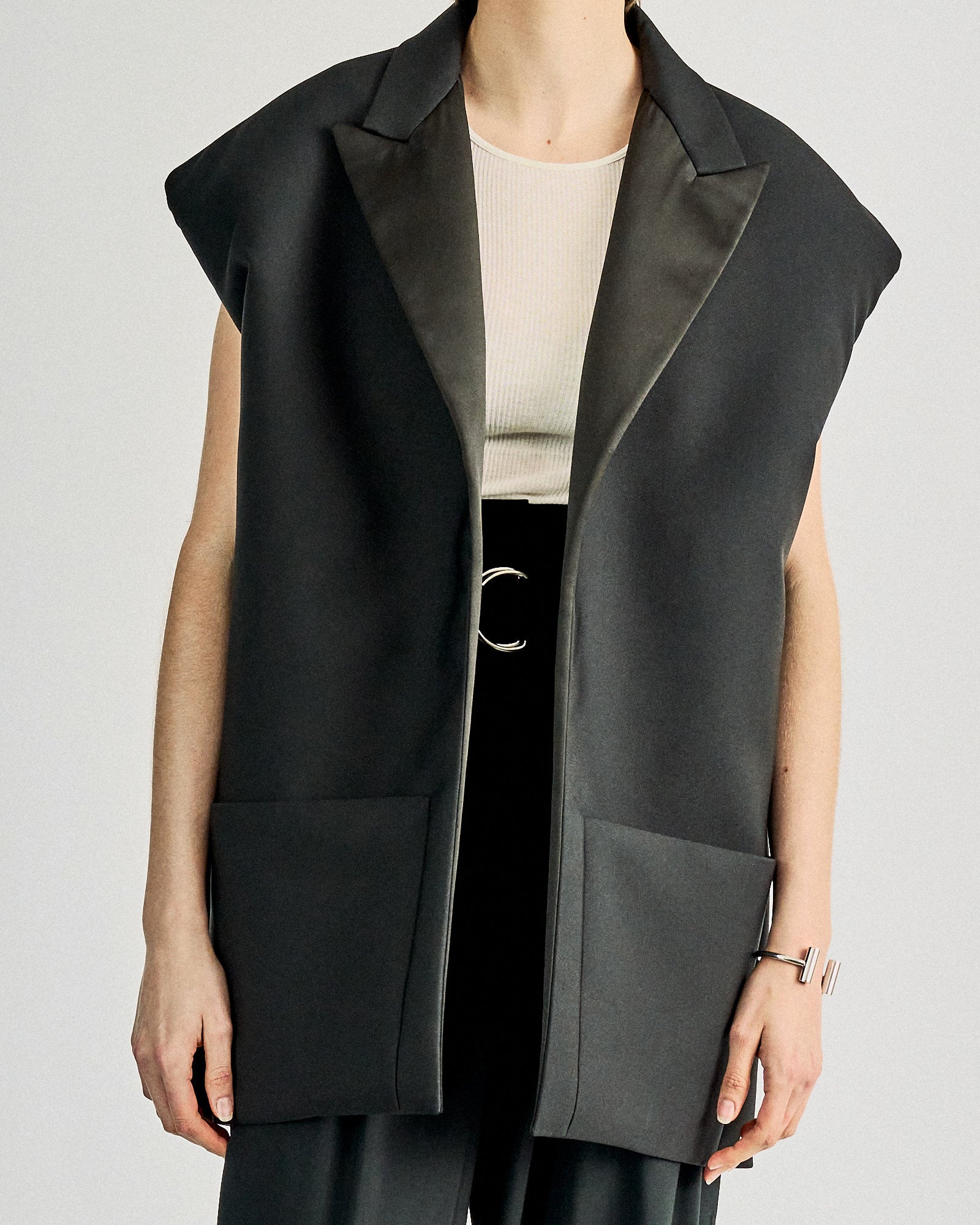 A person wearing a sleeveless, oversized black Celine blazer with satin lapels over a white ribbed top and dark pants, hands relaxed at sides.