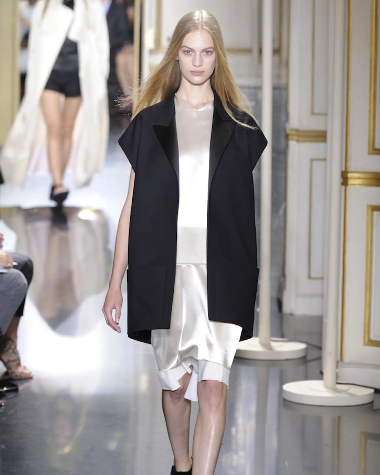 Model walking on a runway wearing a black Celine coat over a white dress.
