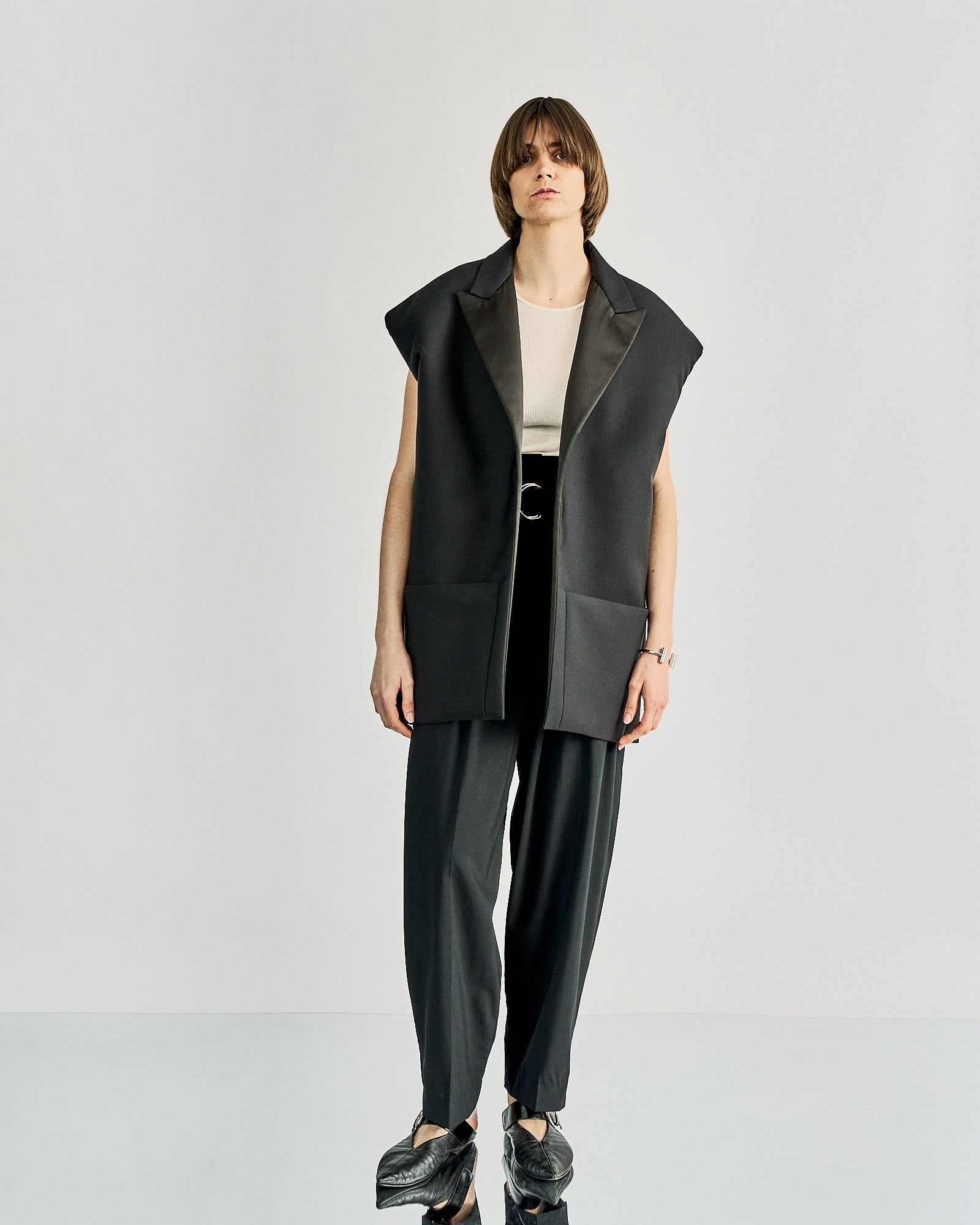 A person wearing a sleeveless, oversized black Celine blazer with satin lapels over a white ribbed top and dark pants, hands relaxed at sides.