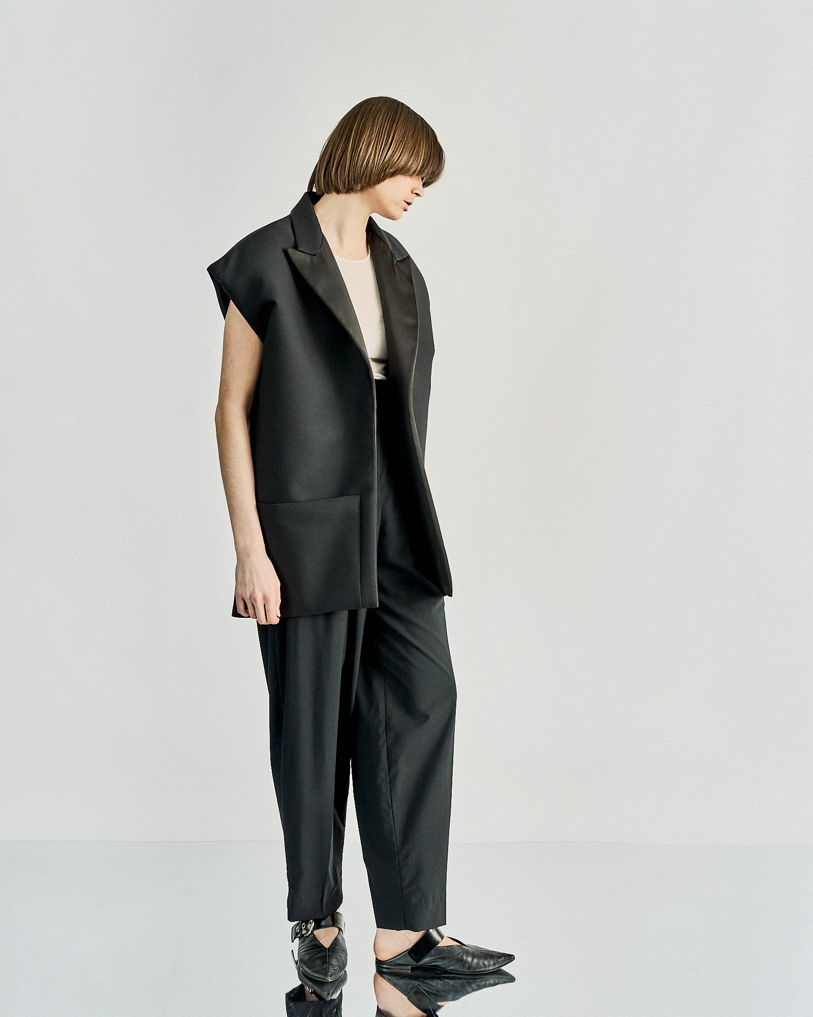 A person wearing a sleeveless, oversized black Celine blazer with satin lapels over a white ribbed top and dark pants, hands relaxed at sides.