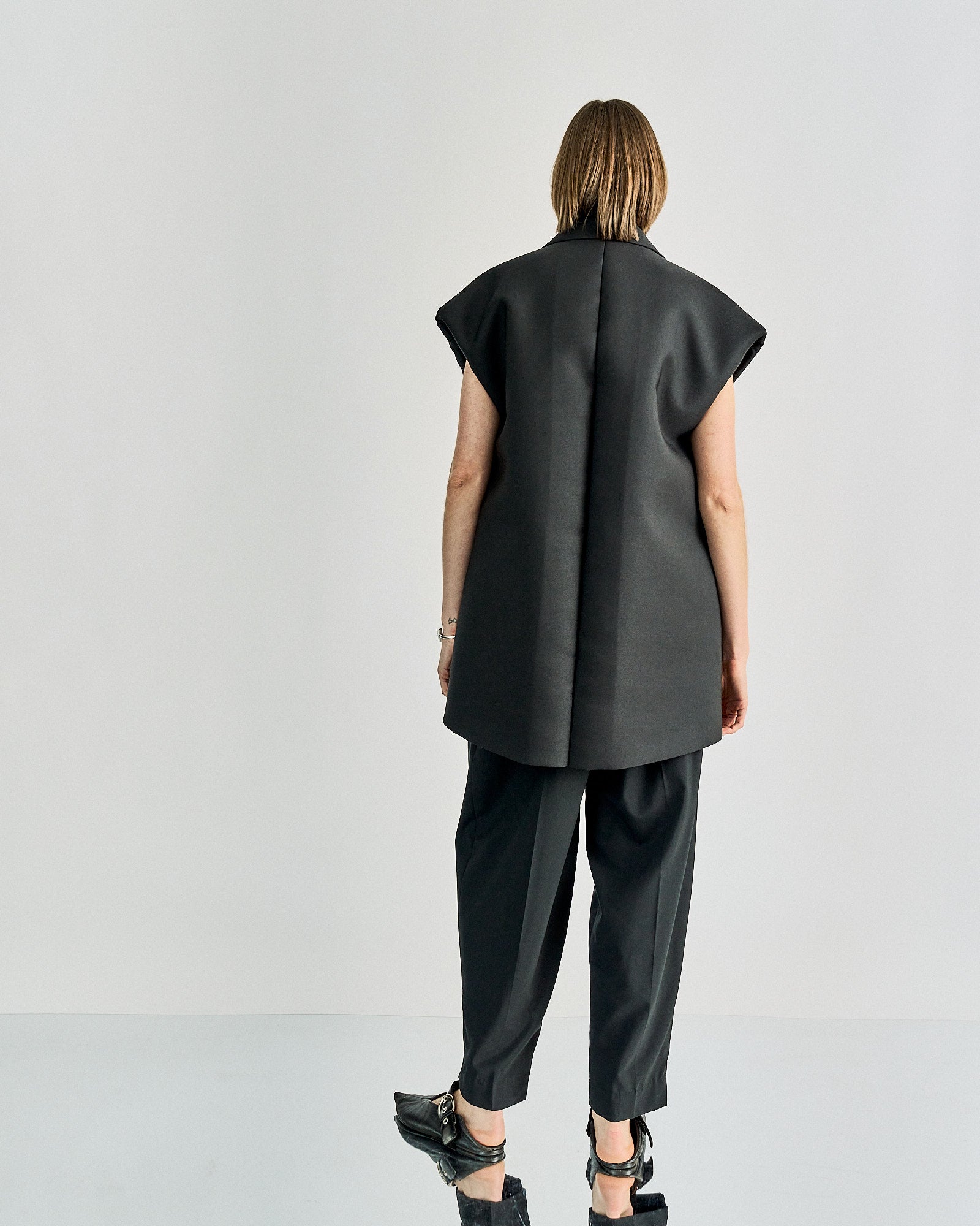 A person stands facing away, wearing a sleeveless black Celine blazer and matching pants. The outfit conveys a modern, minimalist, and professional tone.