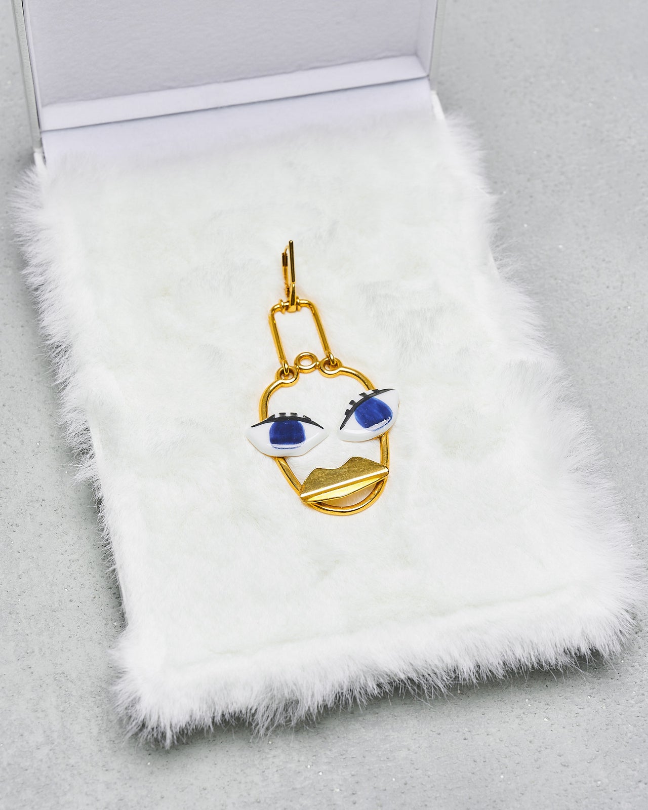 A unique gold-coloured Celine earring with a face design featuring bold blue eyes, presented on a white faux fur surface.
