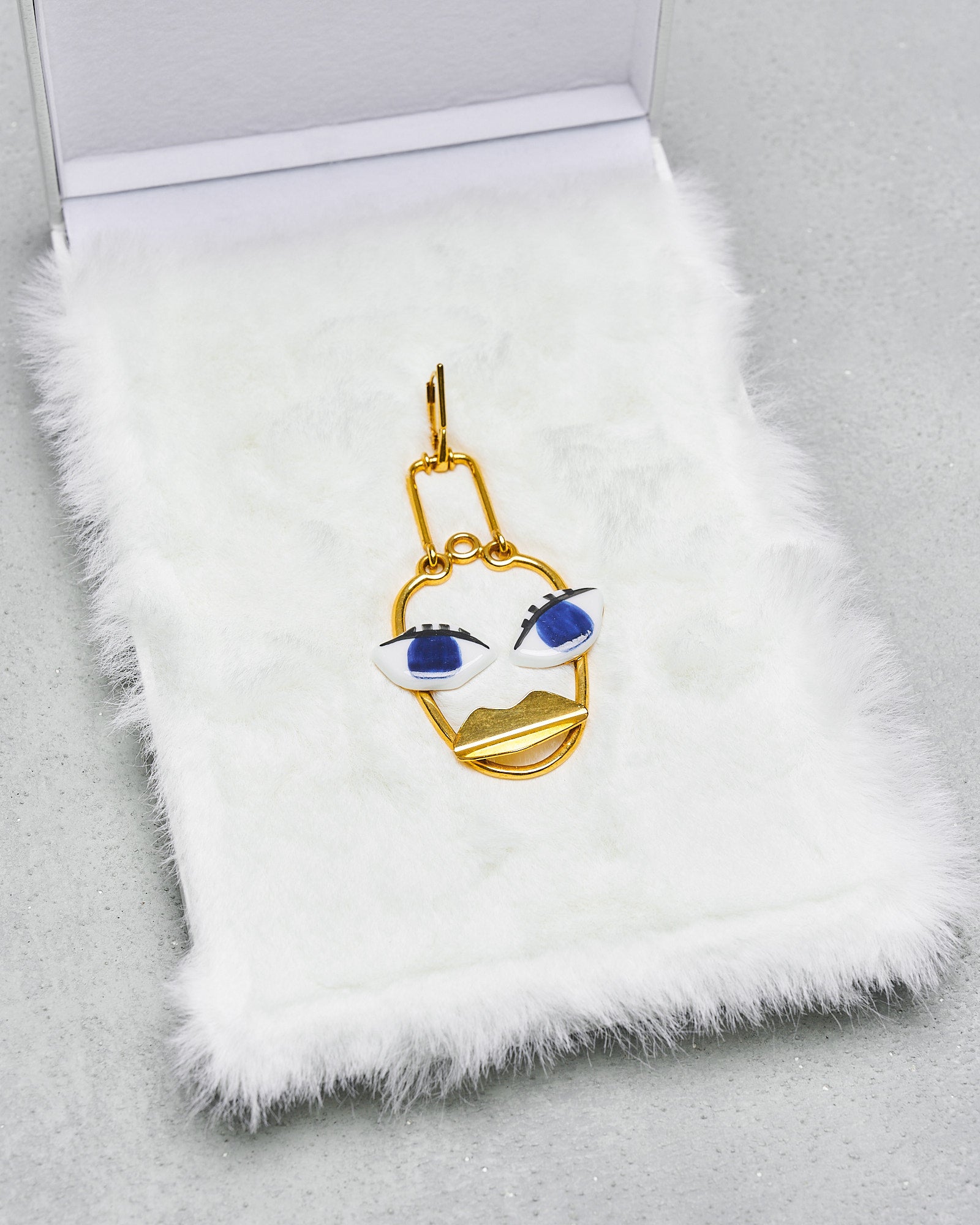 A unique gold-coloured Celine earring with a face design featuring bold blue eyes, presented on a white faux fur surface.