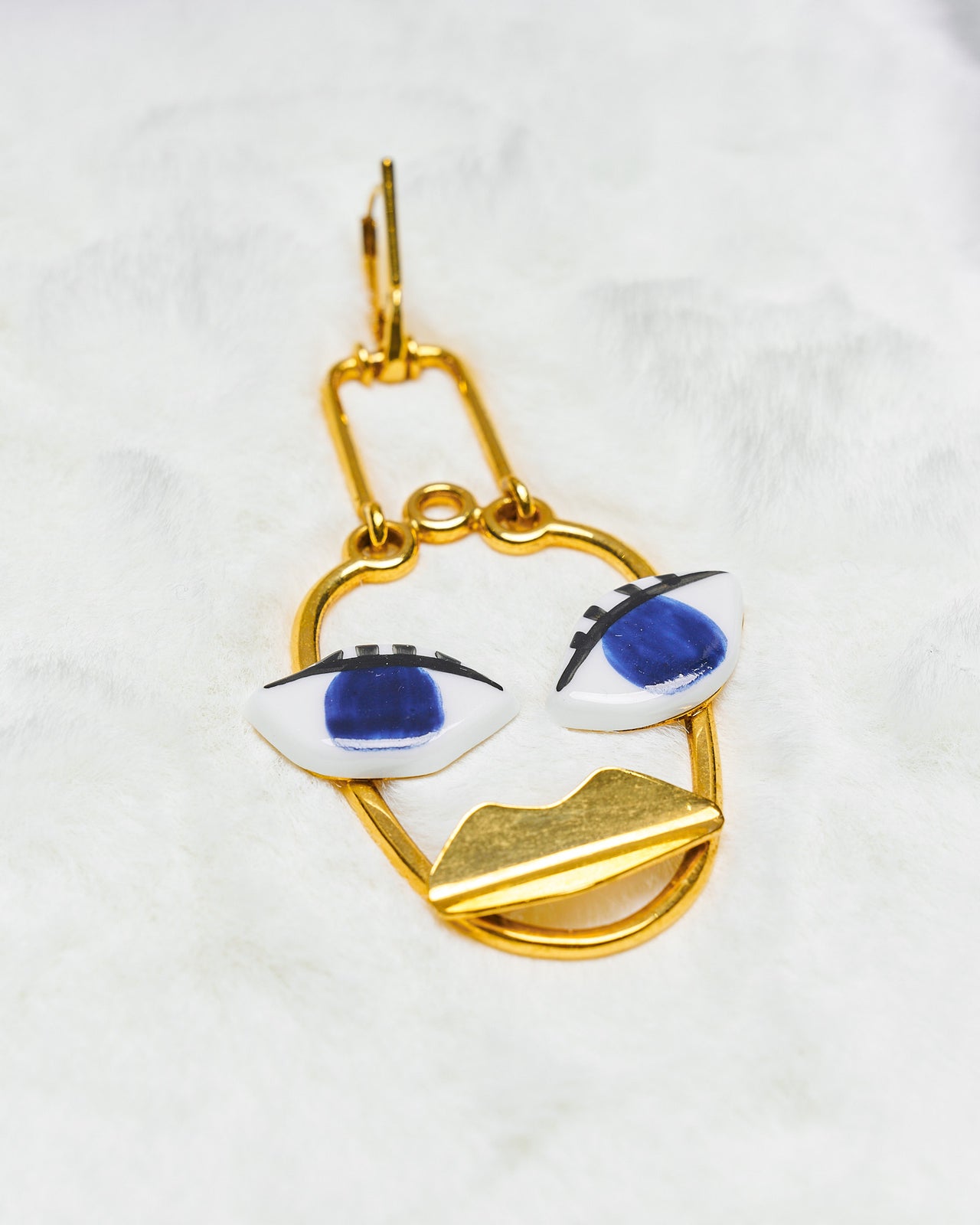A unique gold-coloured Celine earring with a face design featuring bold blue eyes, presented on a white faux fur surface.