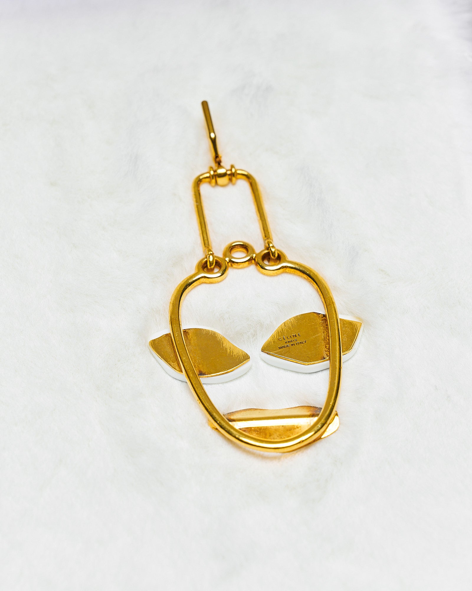 A minimalist gold Celine earring is shaped like an abstract face with a smooth, open oval for a head, angular features for eyes, and a subtle smile on a white background