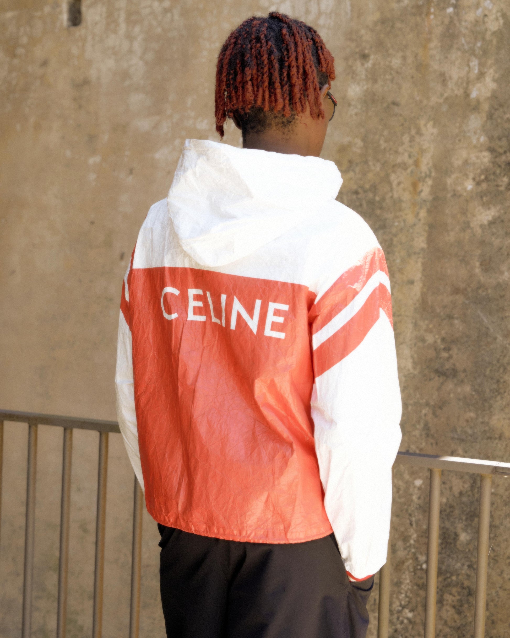 Person in a red and white Celine windbreaker with braided hair faces a textured stone wall. The mood is casual and stylish.