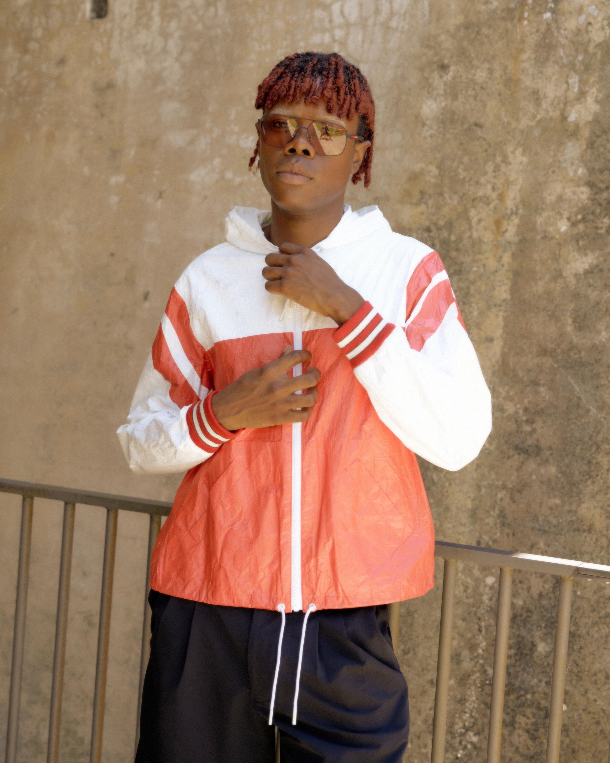 Young person with braided red hair wearing sunglasses and a red and white Celine jacket. They stand confidently against a textured wall, conveying a casual, stylish vibe.
