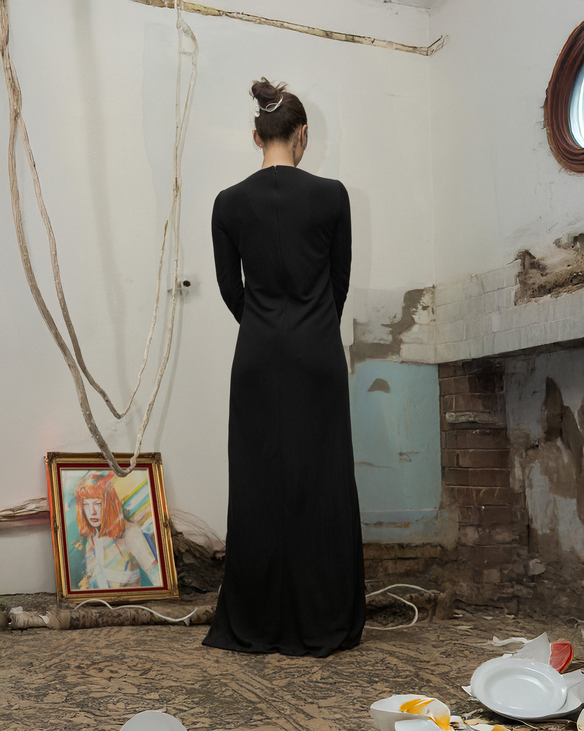 Person wearing a long black Celine dress standing in a room with peeling paint and a painting on the wall.