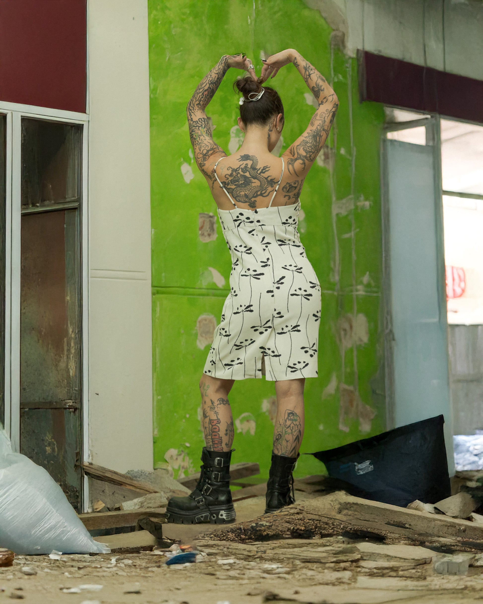 Person with tattoos  wearing a Chanel mini dress standing in an abandoned building with green walls and debris on the floor.