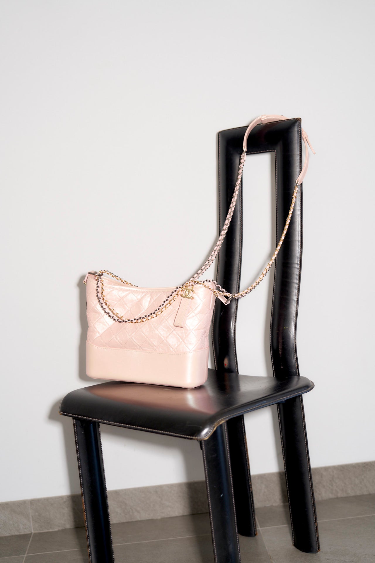 A pale pink quilted Chanel Gabrielle handbag with a chain strap is elegantly draped over a sleek black chair. The setting is minimalist, with a neutral-toned floor and wall, conveying a chic and stylish tone.