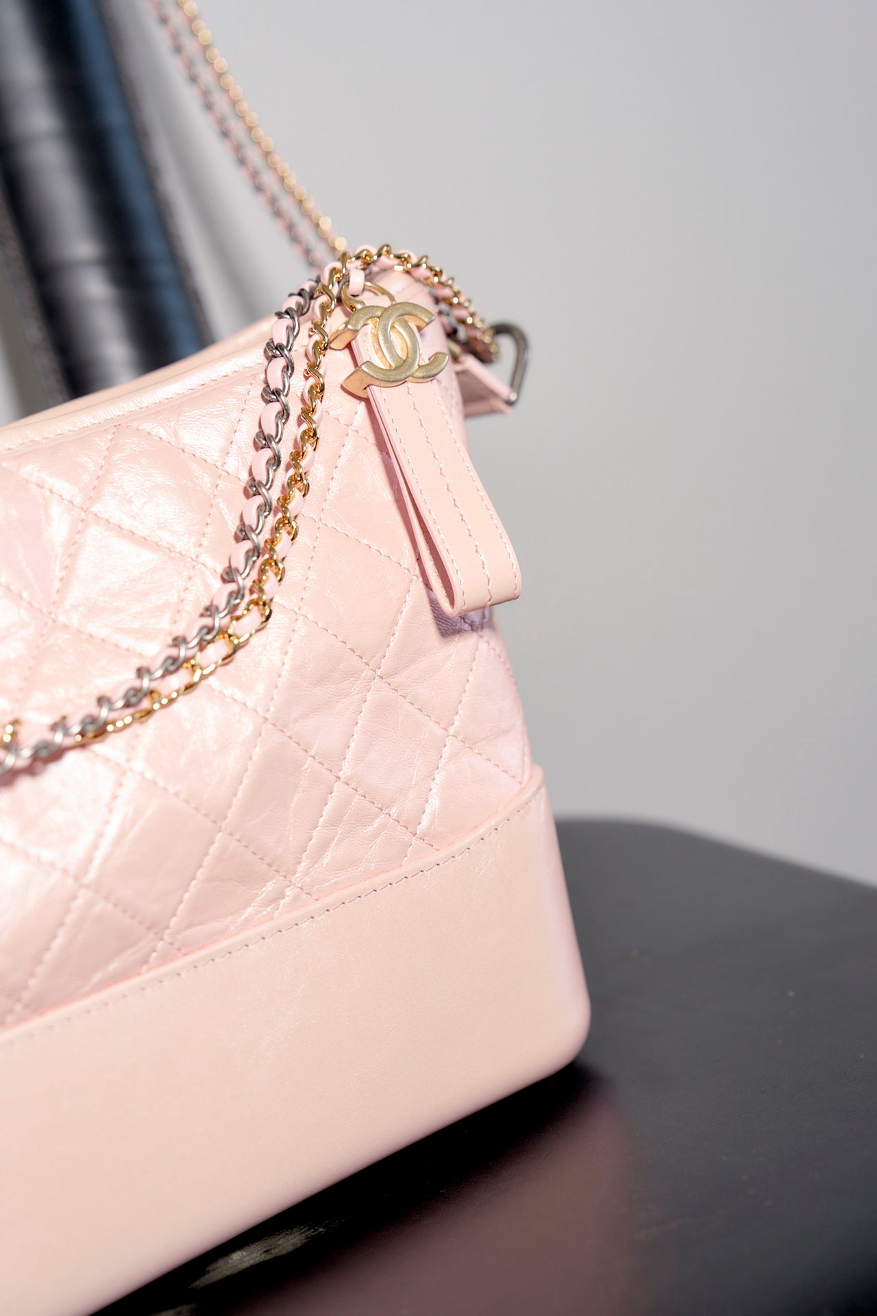 A pale pink quilted Chanel Gabrielle handbag with a chain strap is elegantly draped over a sleek black chair. The setting is minimalist, with a neutral-toned floor and wall, conveying a chic and stylish tone.