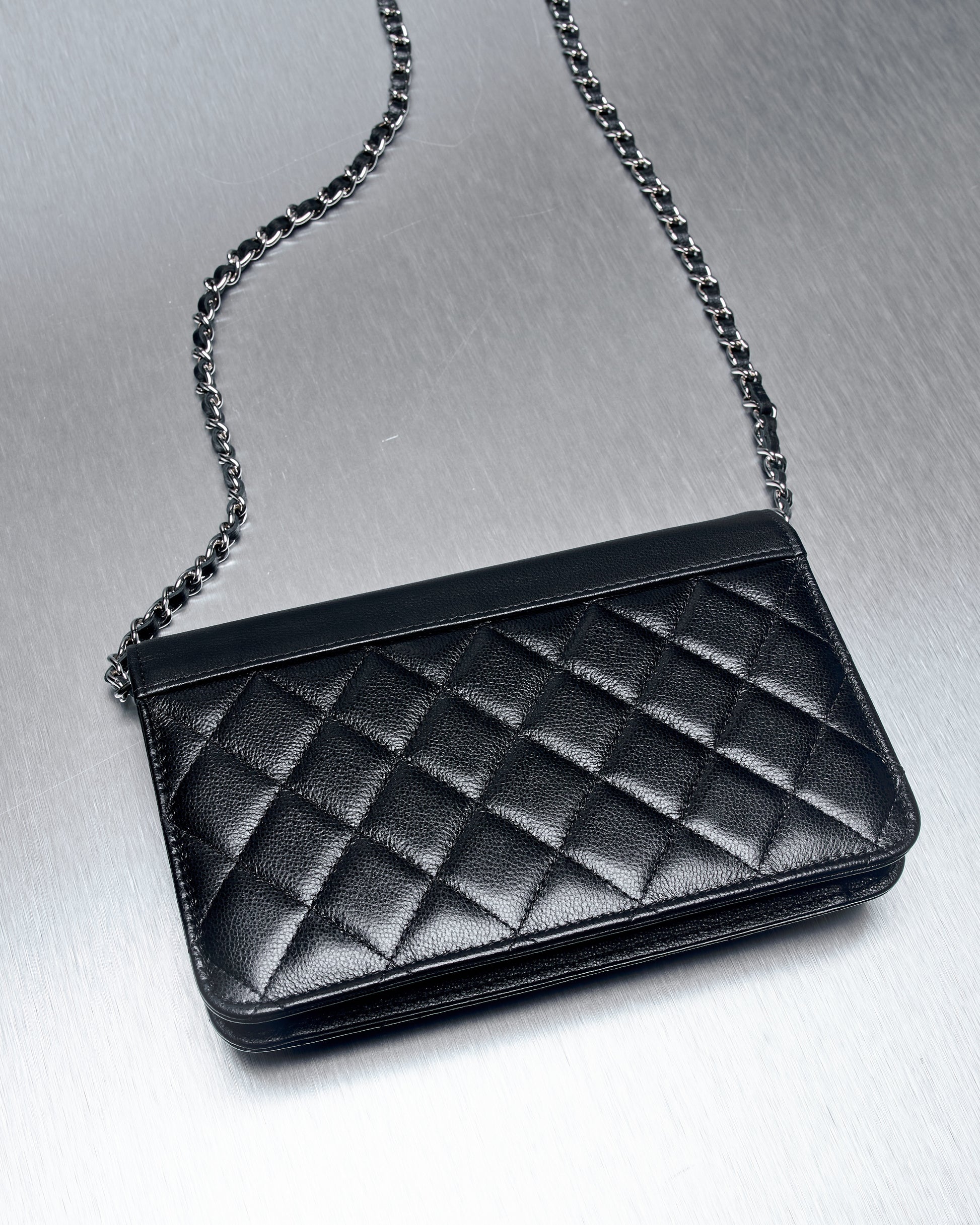 A black leather Chanel handbag with a quilted diamond pattern, featuring a silver chain strap, rests on a brushed metal surface.