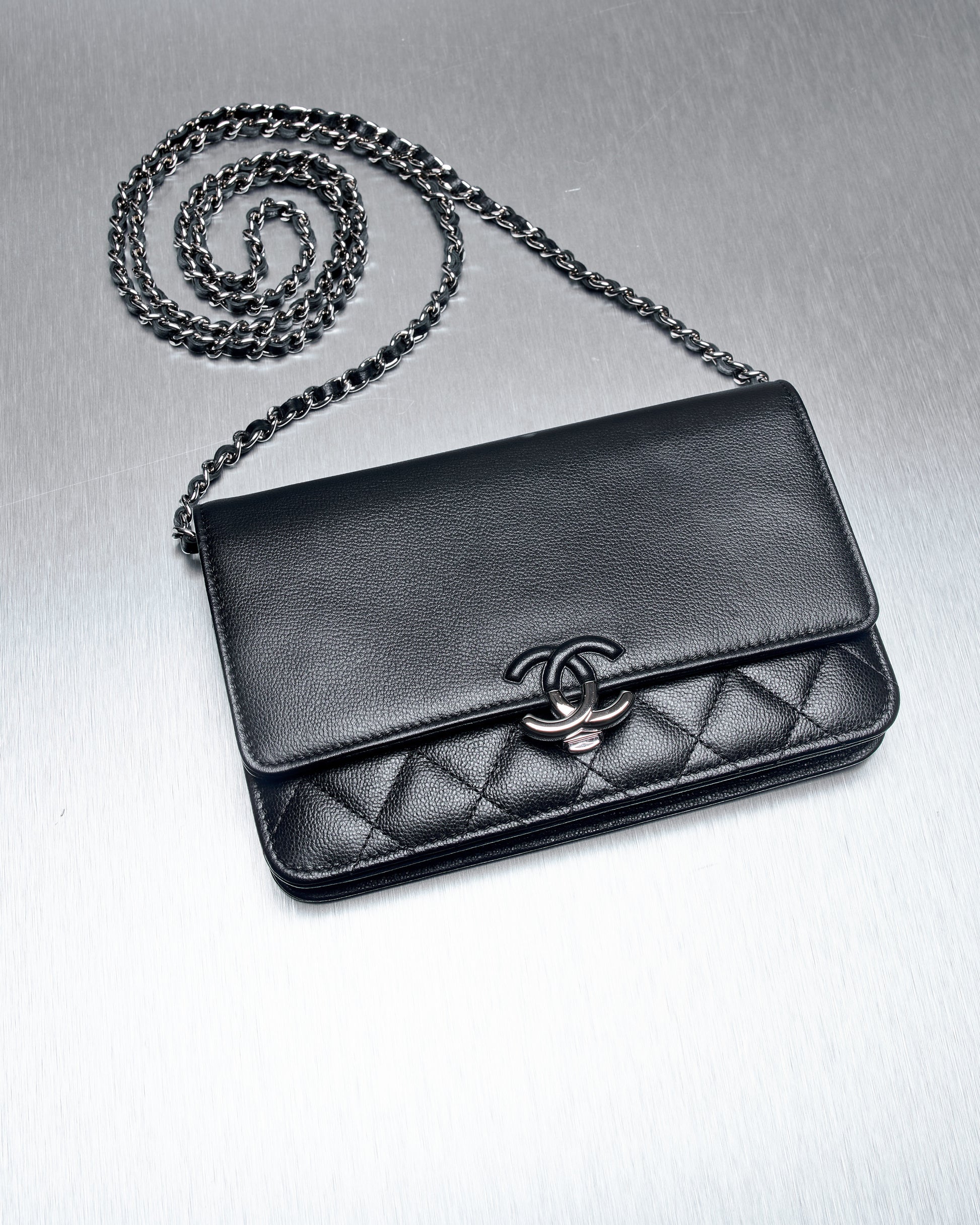 Elegant black leather Chanel handbag with quilted pattern, featuring a silver chain strap and iconic interlocking logo, displayed on a sleek metallic surface.
