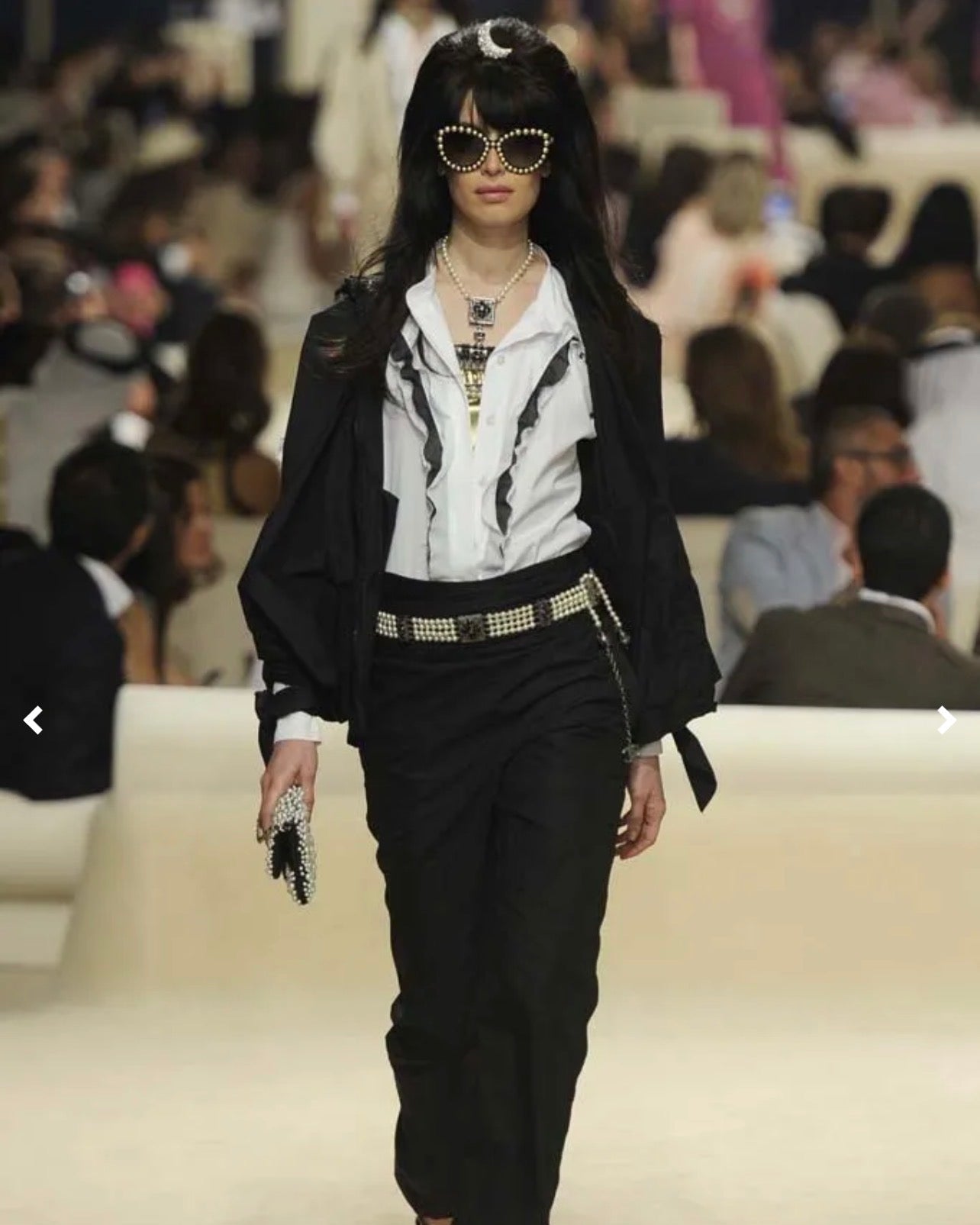 Model walking on a Chanel runway wearing a black outfit with sunglasses, surrounded by an audience.