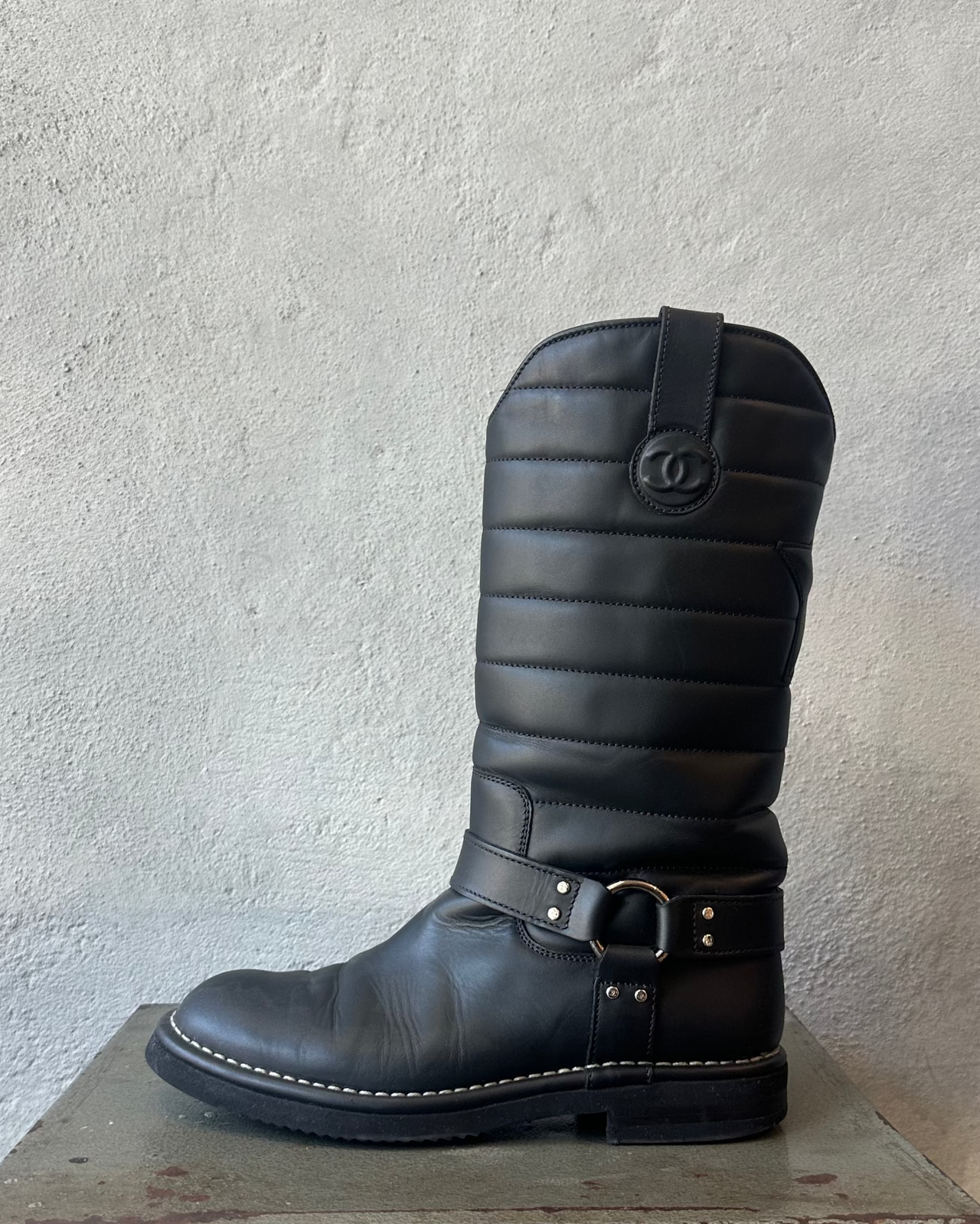 Black quilted Chanel boot with a brand logo on a metal stool against a textured white wall.