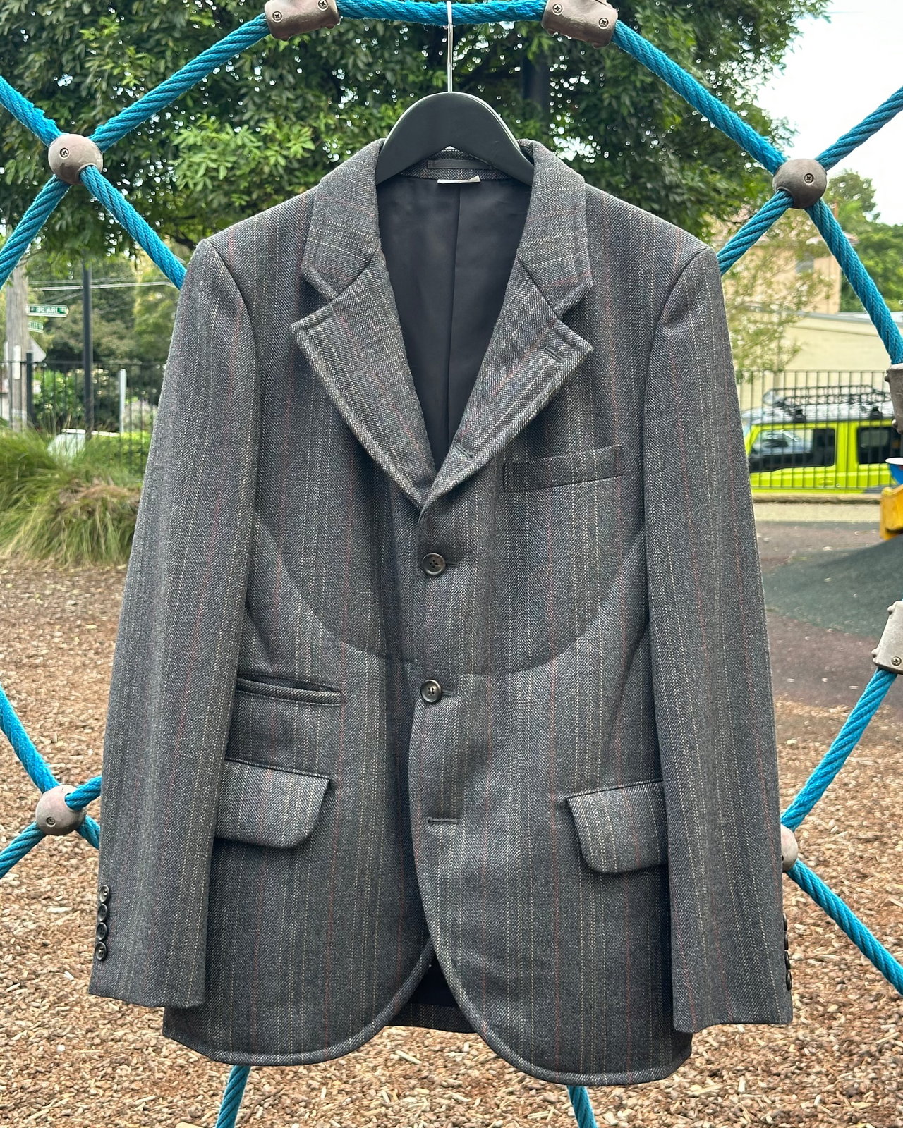 Gray Comme des Garcons Homme Deux blazer on a hanger against a playground background