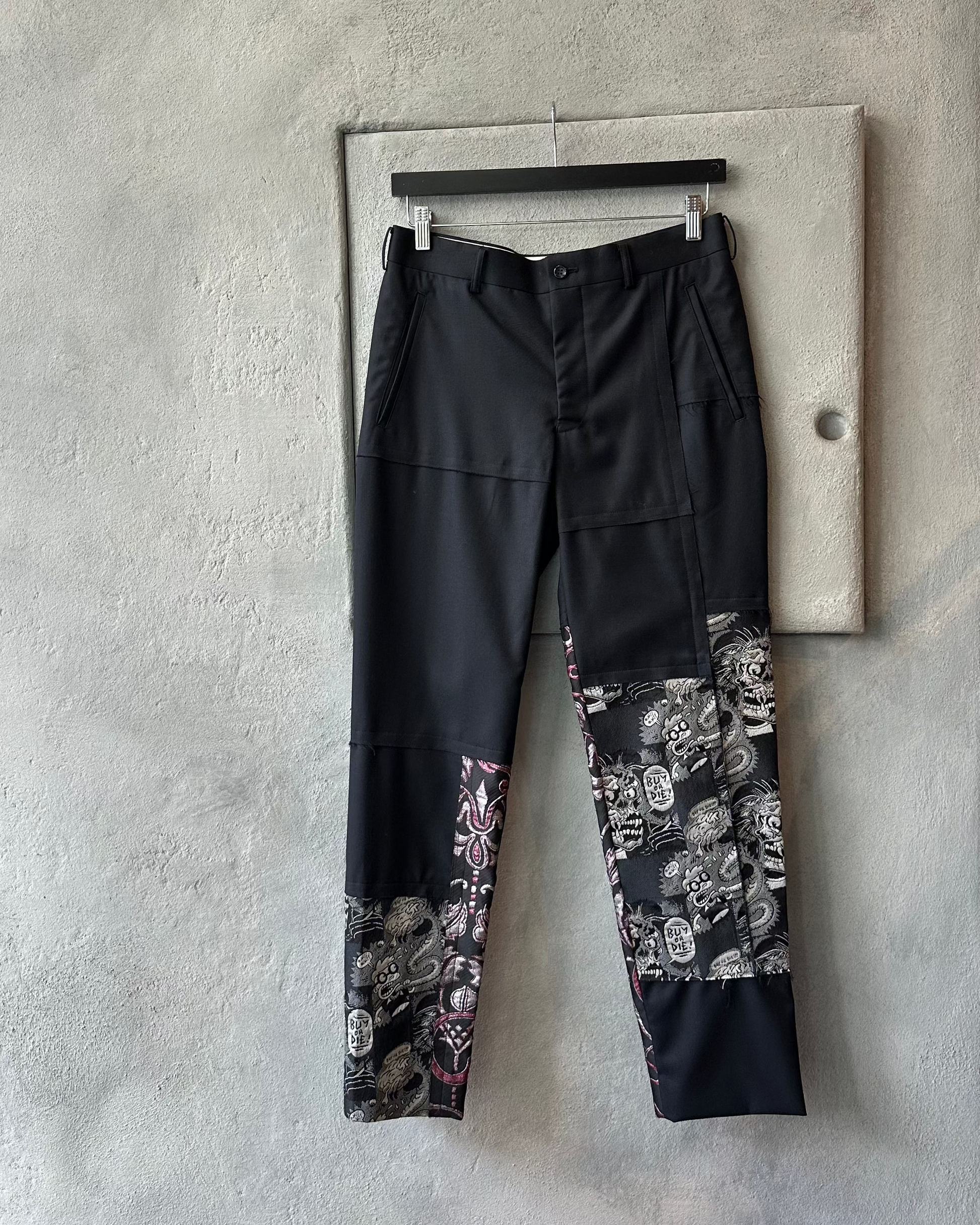 Black Comme des Garcons pants with floral patterns hanging on a concrete wall.