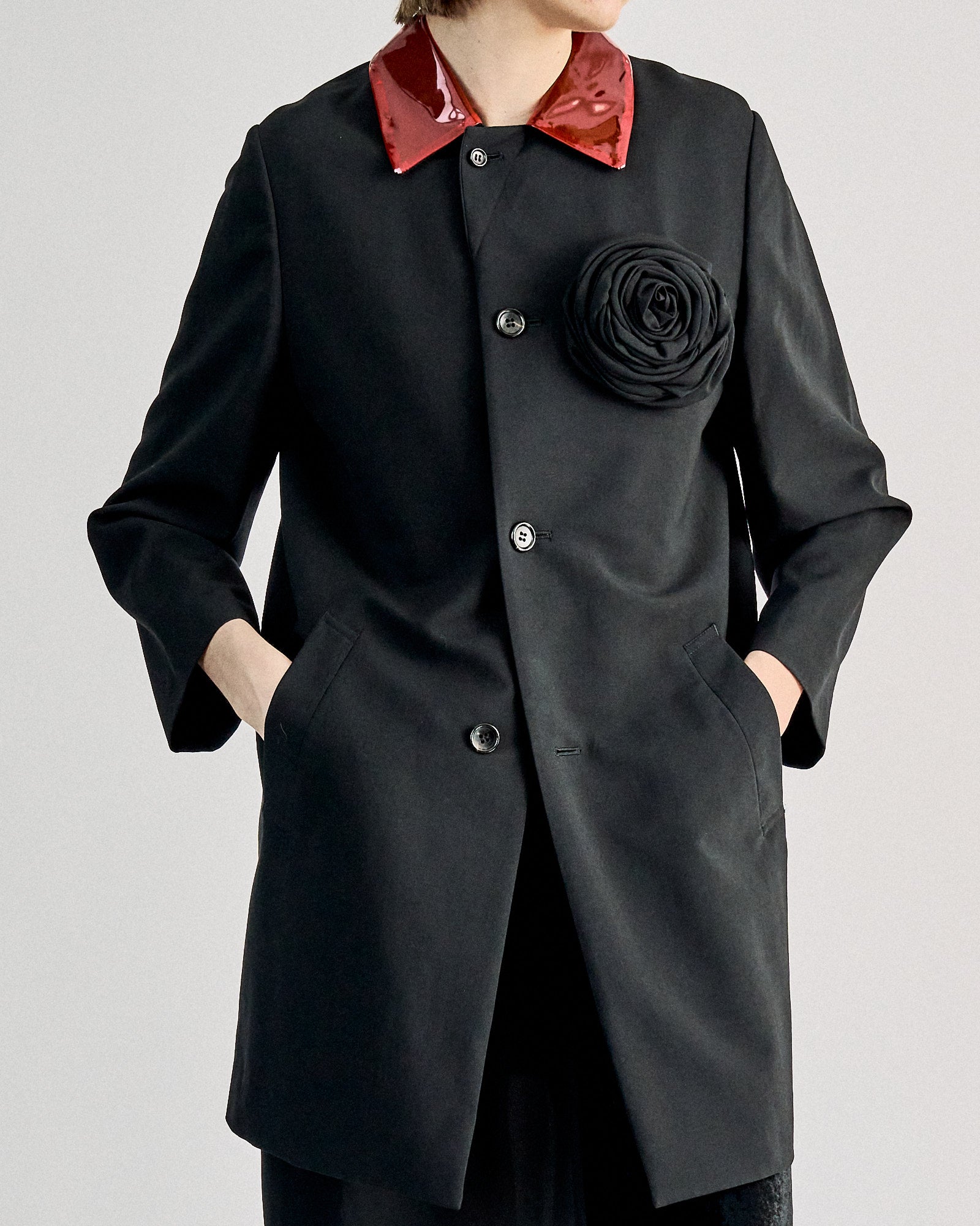 A person wears a chic black Comme des Garcons coat with a glossy red collar and a large black flower on the chest, conveying a sophisticated and stylish tone.