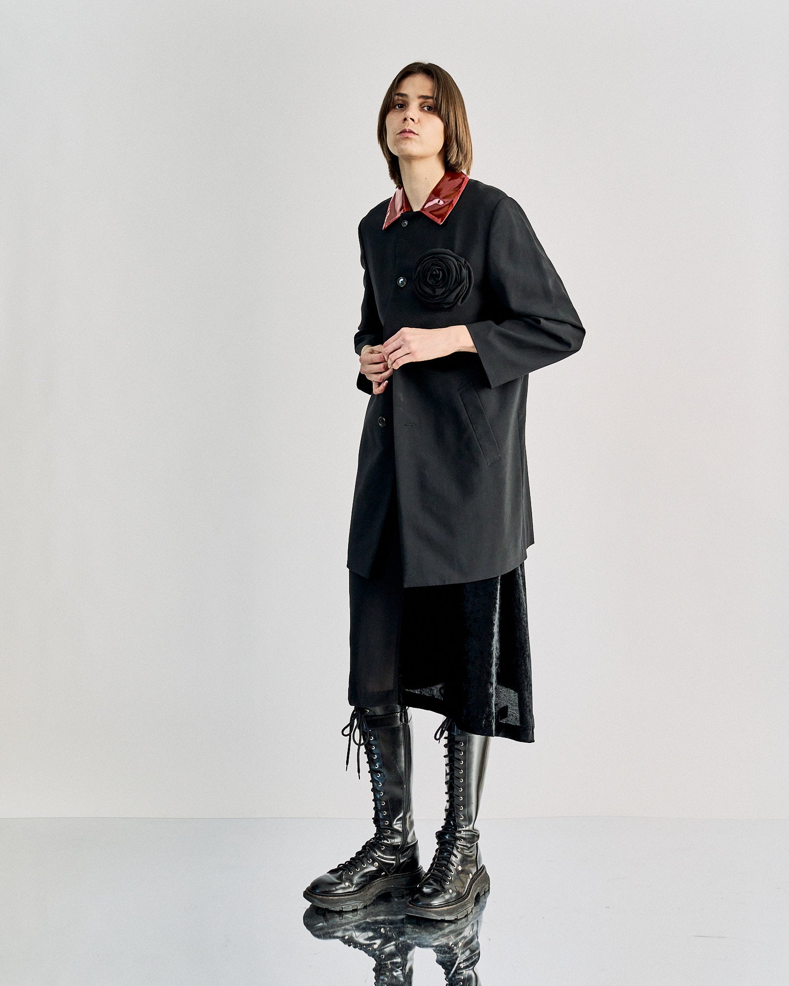 A person wears a chic black Comme des Garcons coat with a glossy red collar and a large black flower on the chest, conveying a sophisticated and stylish tone.