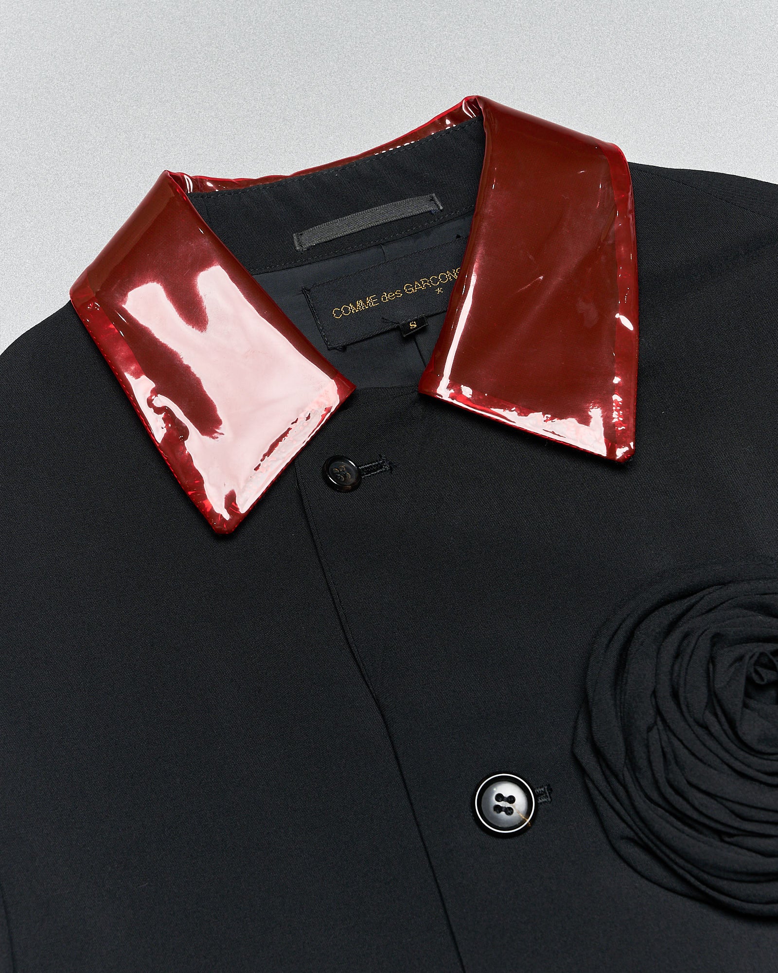 Close-up of a black Comme des Garcons coat with a shiny red patent leather collar. Features classic buttons and a fabric rosette detail, conveying a bold, avant-garde style.