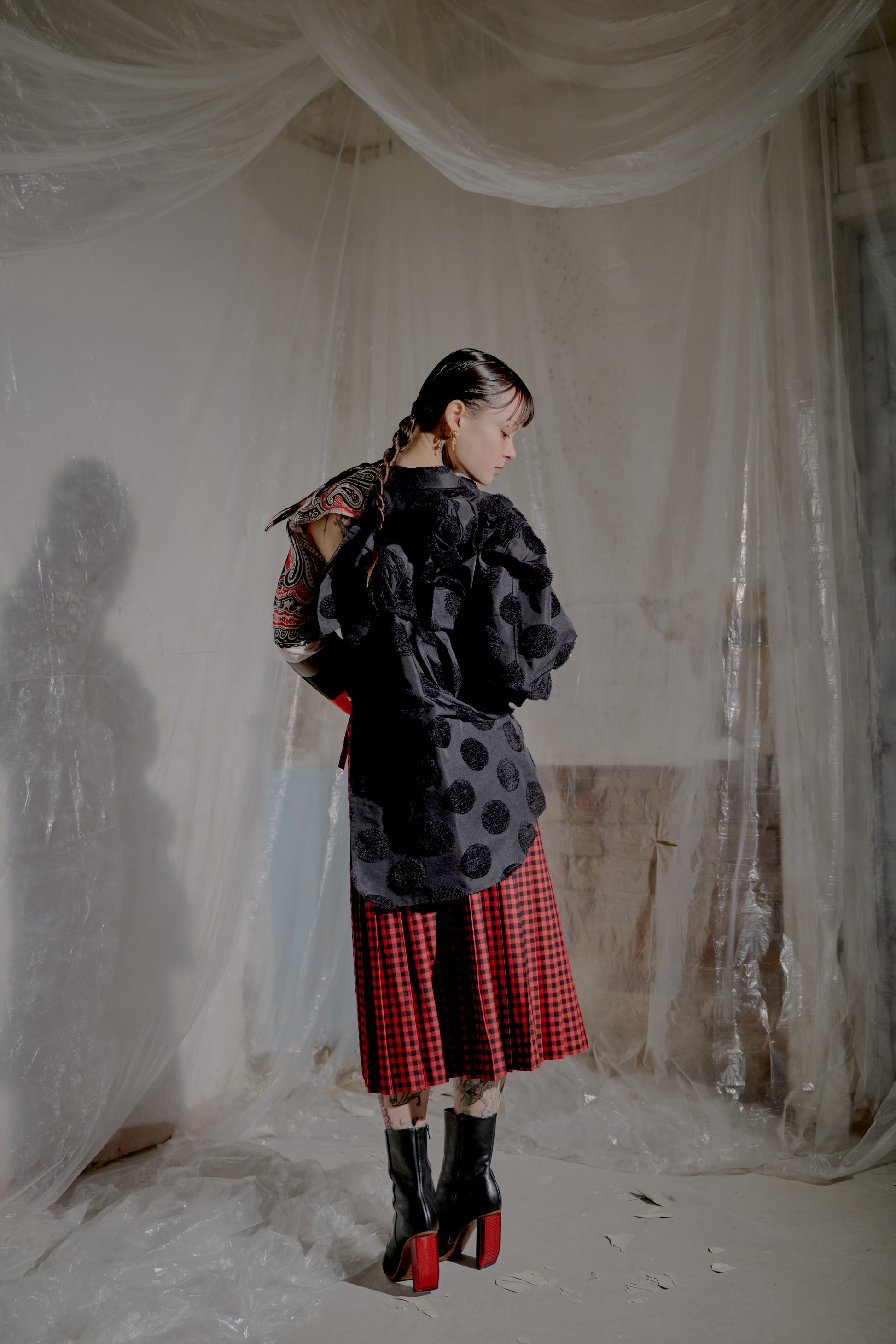 Person wearing a black polka dot Comme Des Garcons coat and red plaid skirt in a room with white curtains.