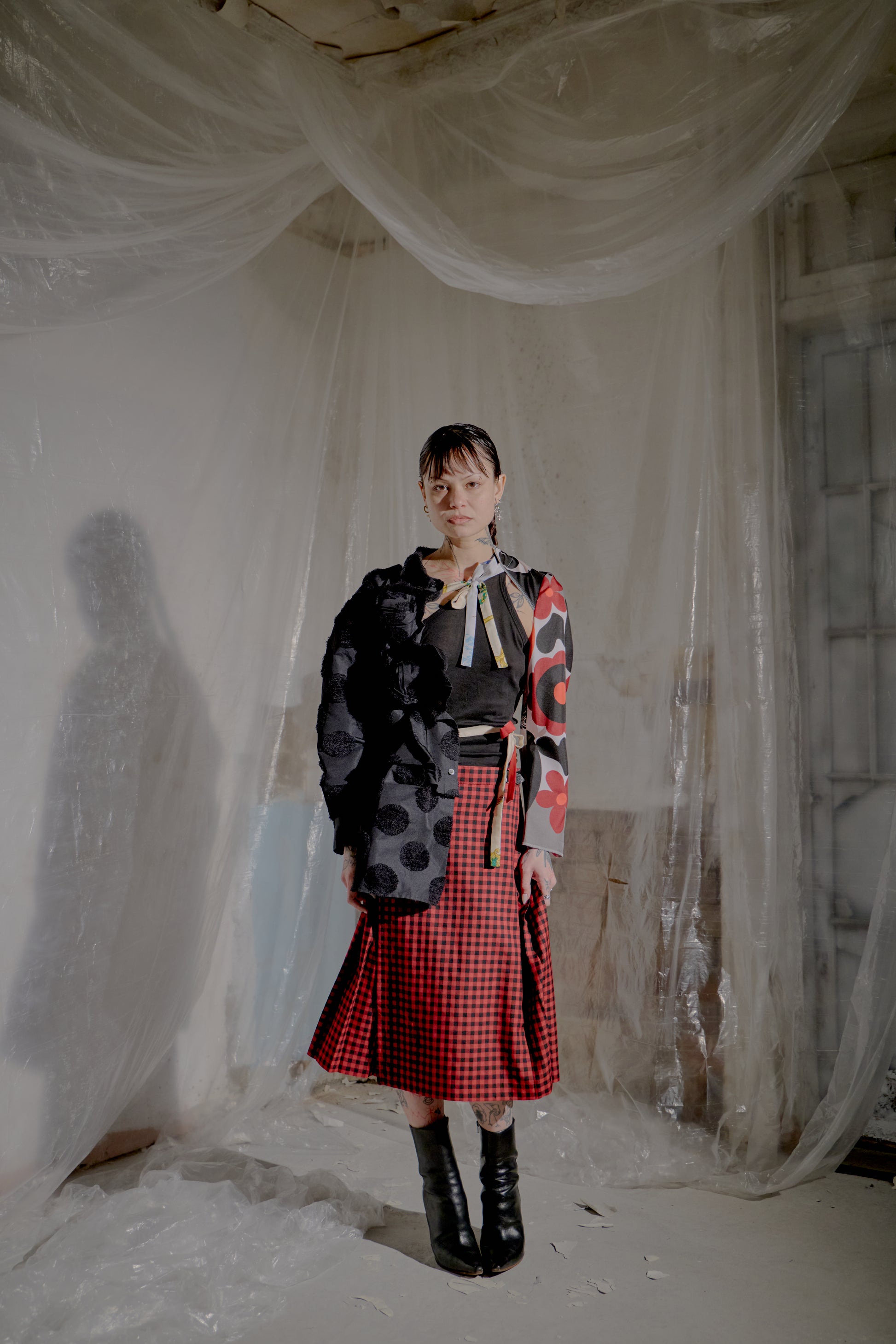 Person wearing a black and red Comme Des Garcons outfit with a checkered skirt in a room with white fabric.